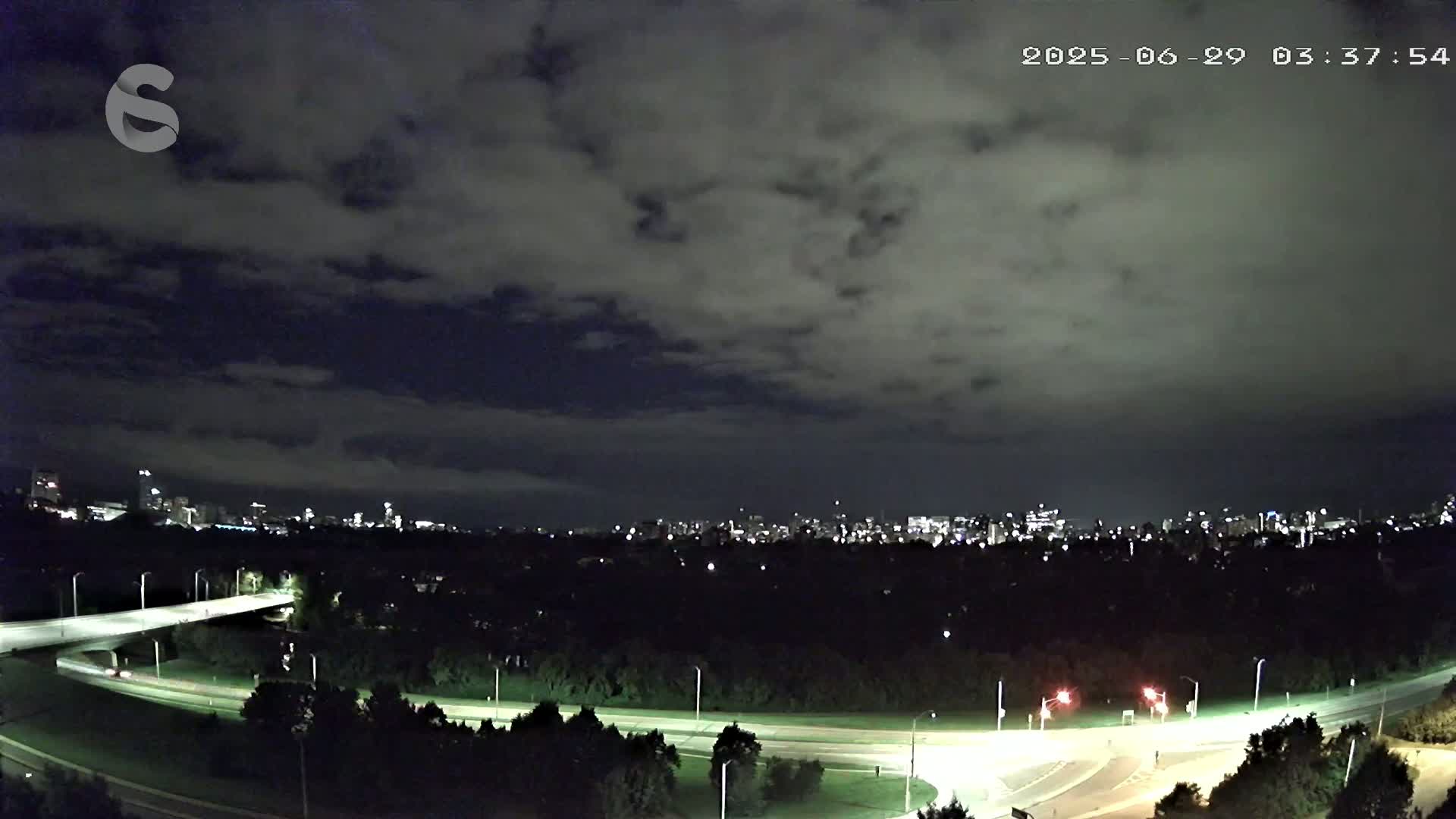 A nighttime view of a city skyline across a dark, tree-covered landscape with a roadway curving through it under a cloudy sky.