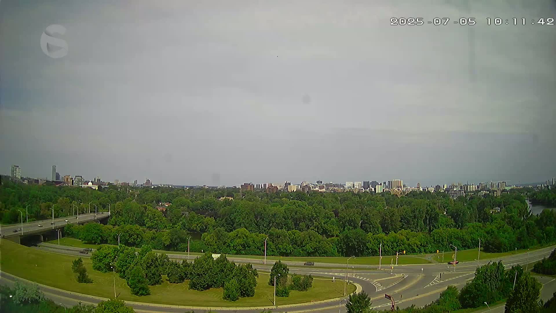 A highway interchange curves through a lush green park, with a city skyline visible in the distance under an overcast sky.