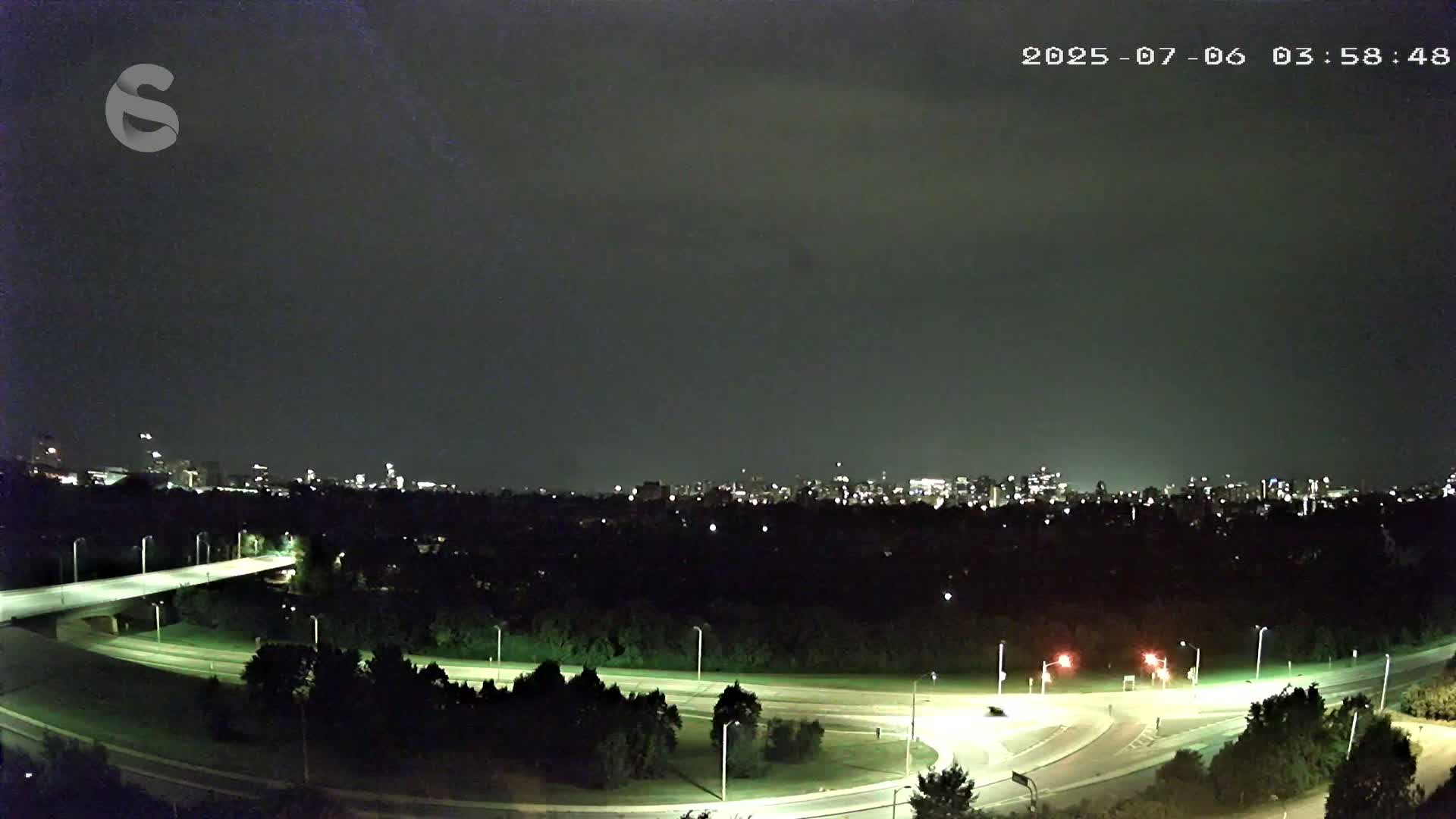 A nighttime view of a city skyline across a tree-lined highway intersection under a dark, cloudy sky.