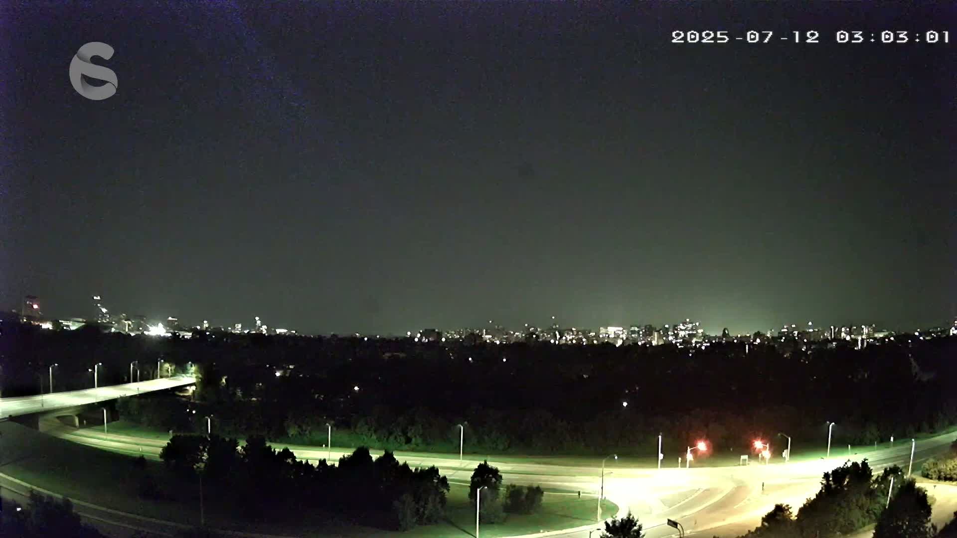A nighttime view of a city skyline across a dark, tree-lined roadway with a few illuminated vehicles.