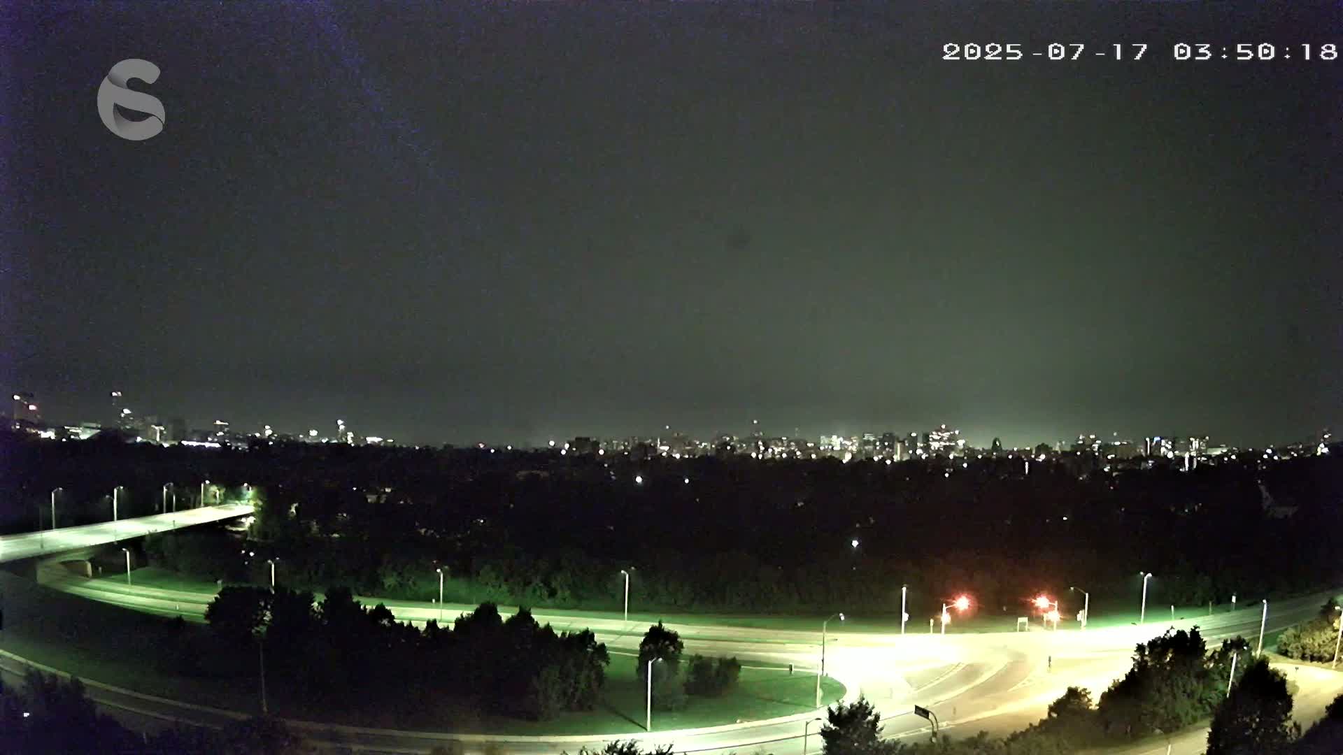 A nighttime view of a city skyline across a dark, tree-lined highway interchange under a cloudy sky.