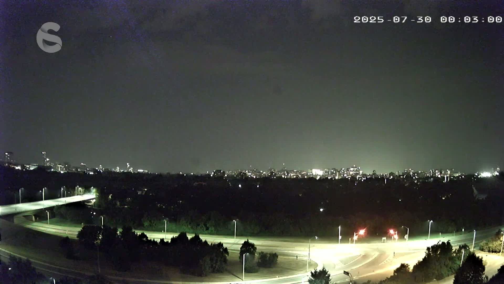 A nighttime view of a city skyline across a dark, tree-lined highway interchange under a cloudy sky.