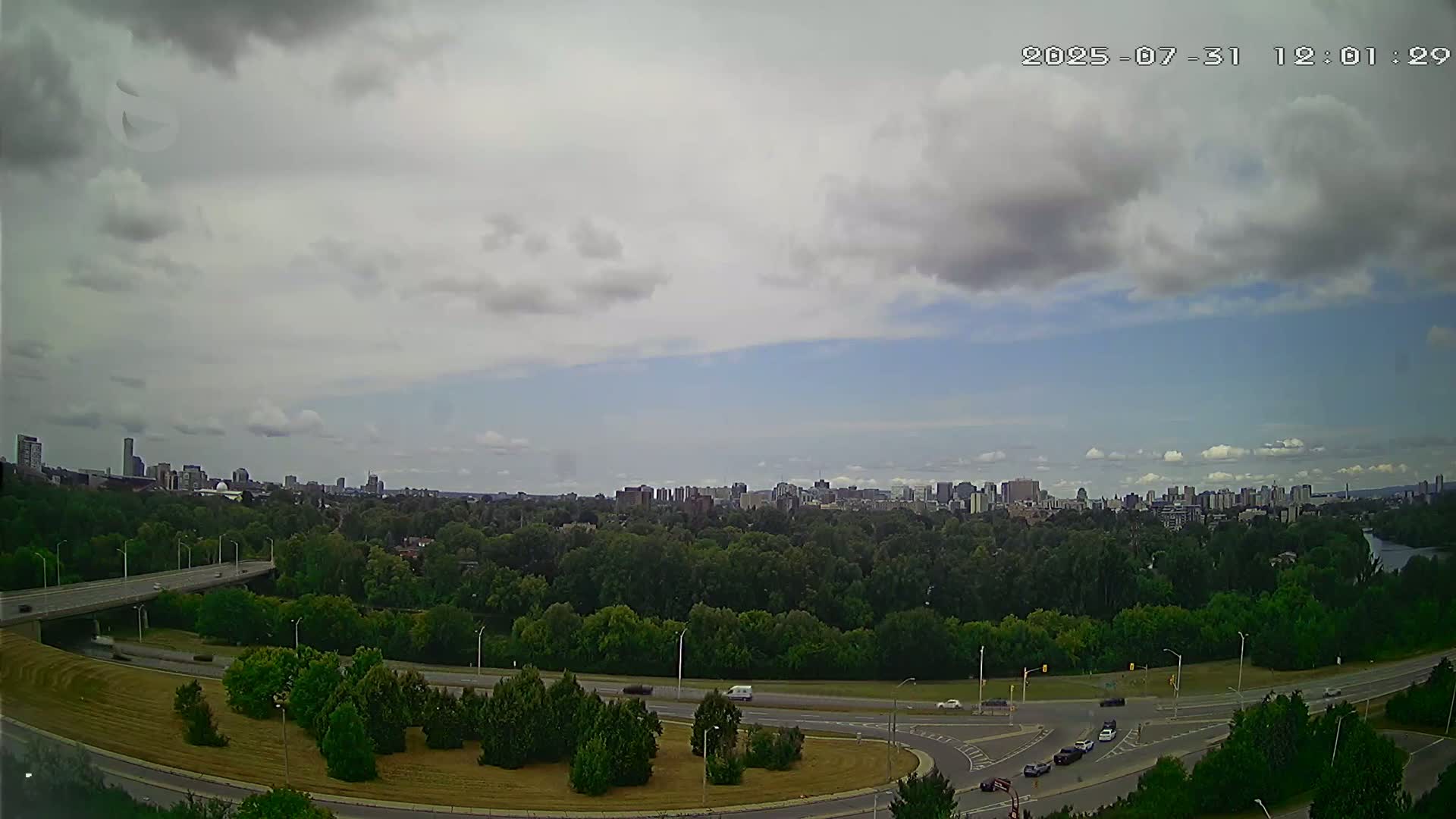 A mostly cloudy sky overlooks a highway curving through a green landscape, leading to a distant city skyline.