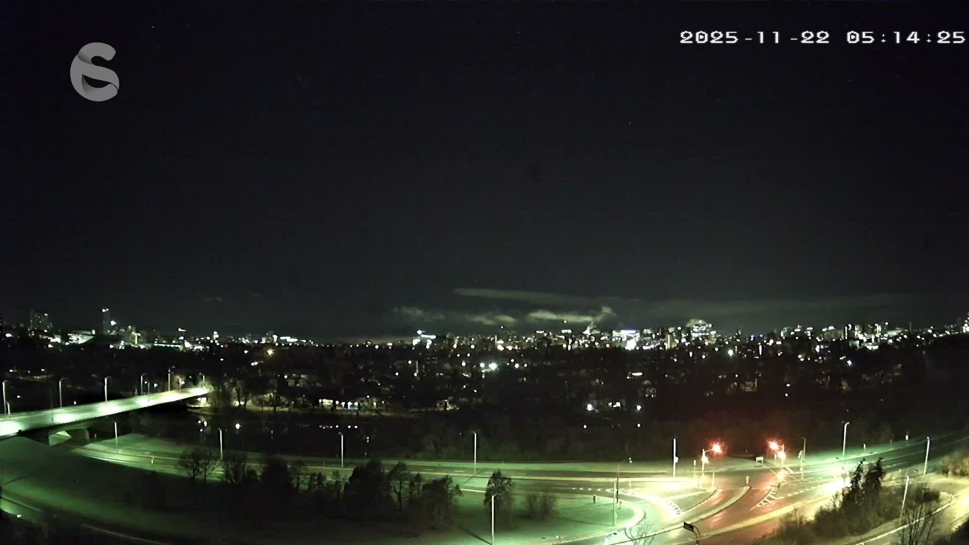 The image shows a nighttime panoramic view of a city skyline with numerous illuminated buildings, a brightly lit bridge, and complex roadways in the foreground, all under a dark, partly cloudy sky.