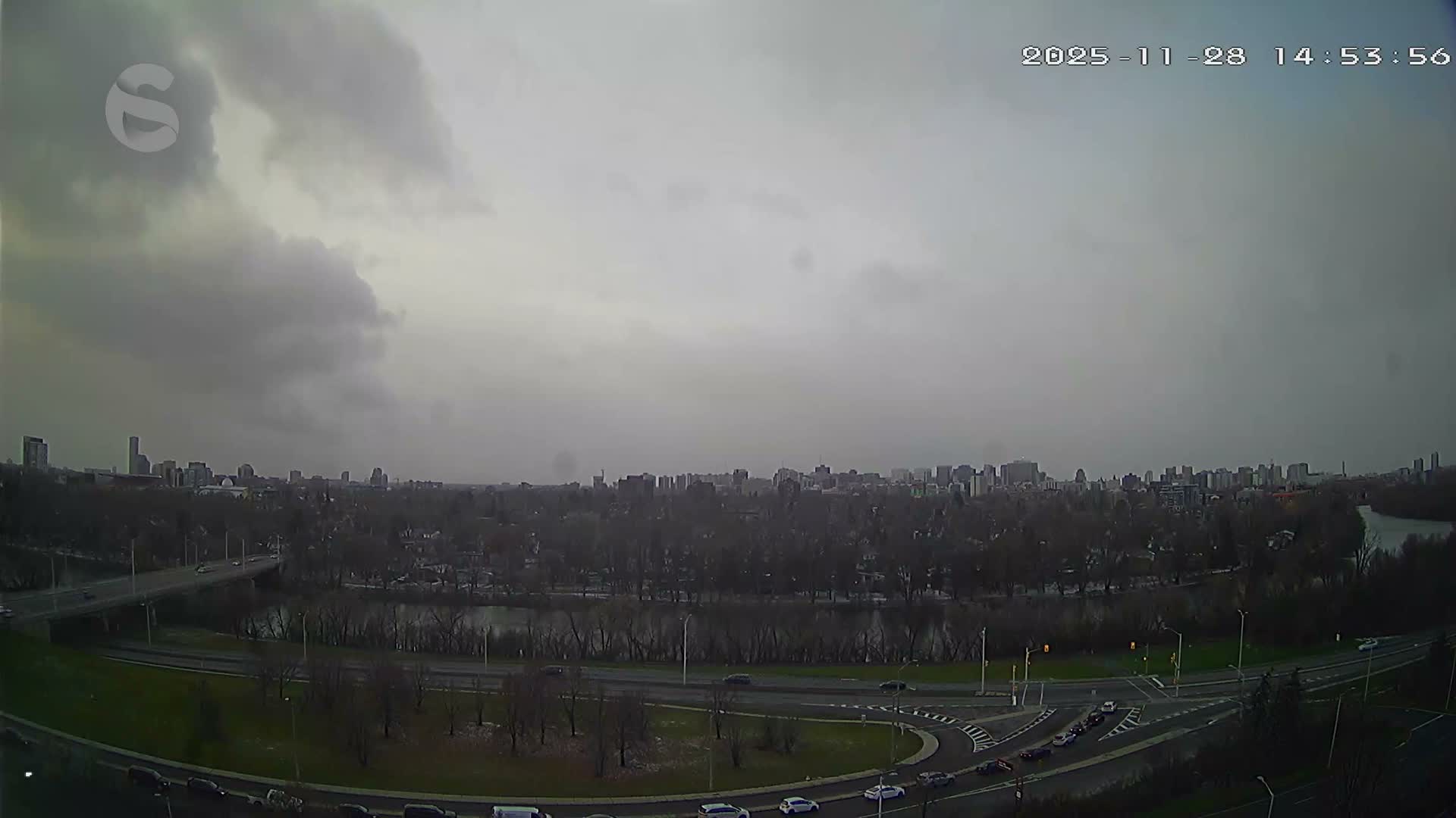 An elevated view reveals an urban landscape under a heavily overcast and grey sky, with a distant city skyline, a wide river bordered by bare trees, and multiple roads with moving traffic in the foreground.