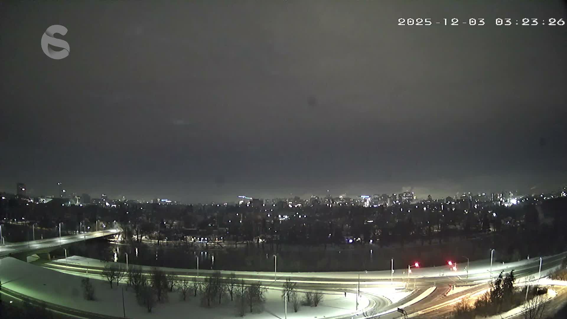 A wide panoramic view captures a snowy cityscape at night, featuring a river, illuminated bridges and roads, and distant city lights under an overcast sky.