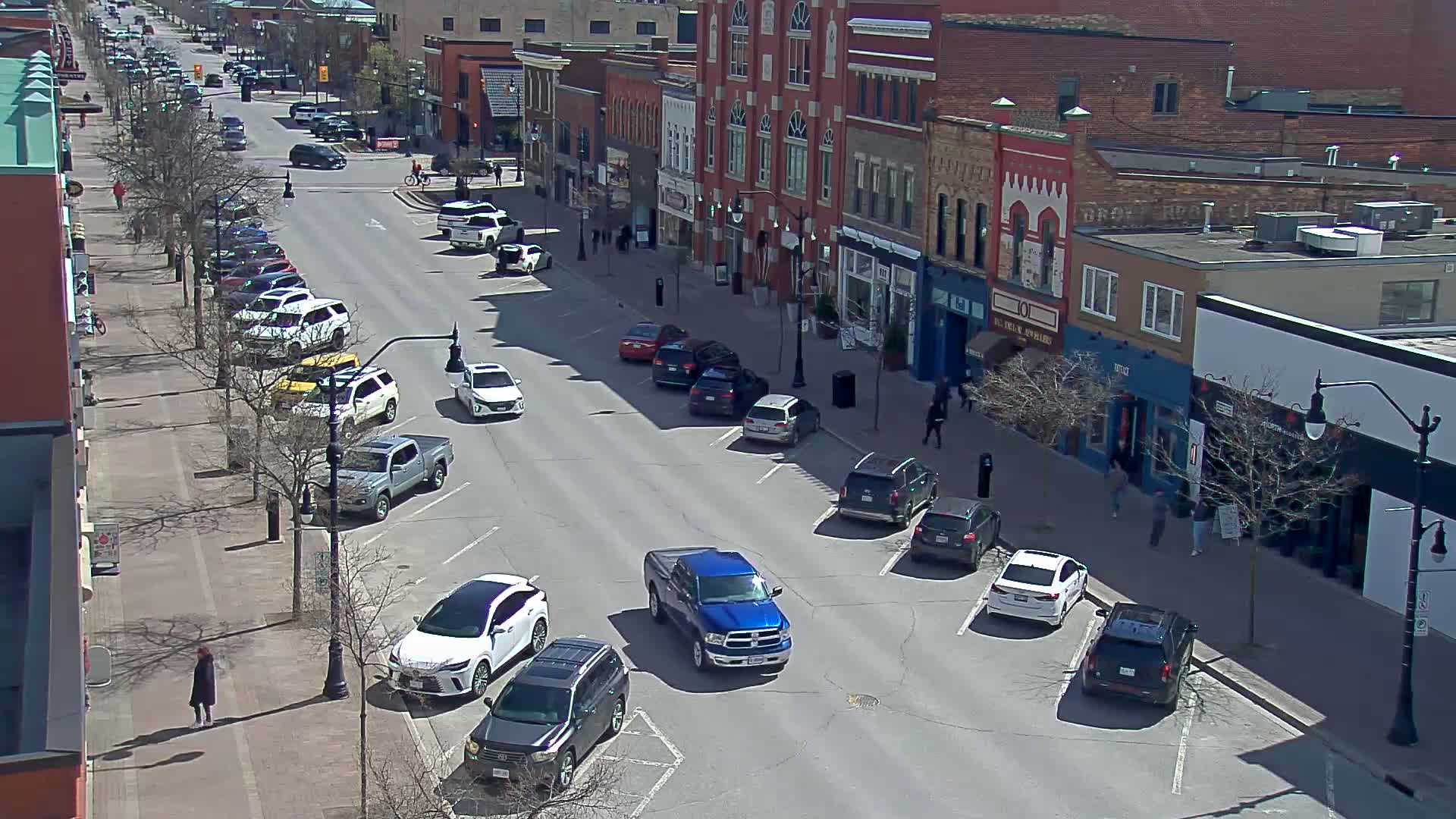 A sunny day reveals a street lined with parked cars and brick buildings, with a few pedestrians walking along the sidewalk.