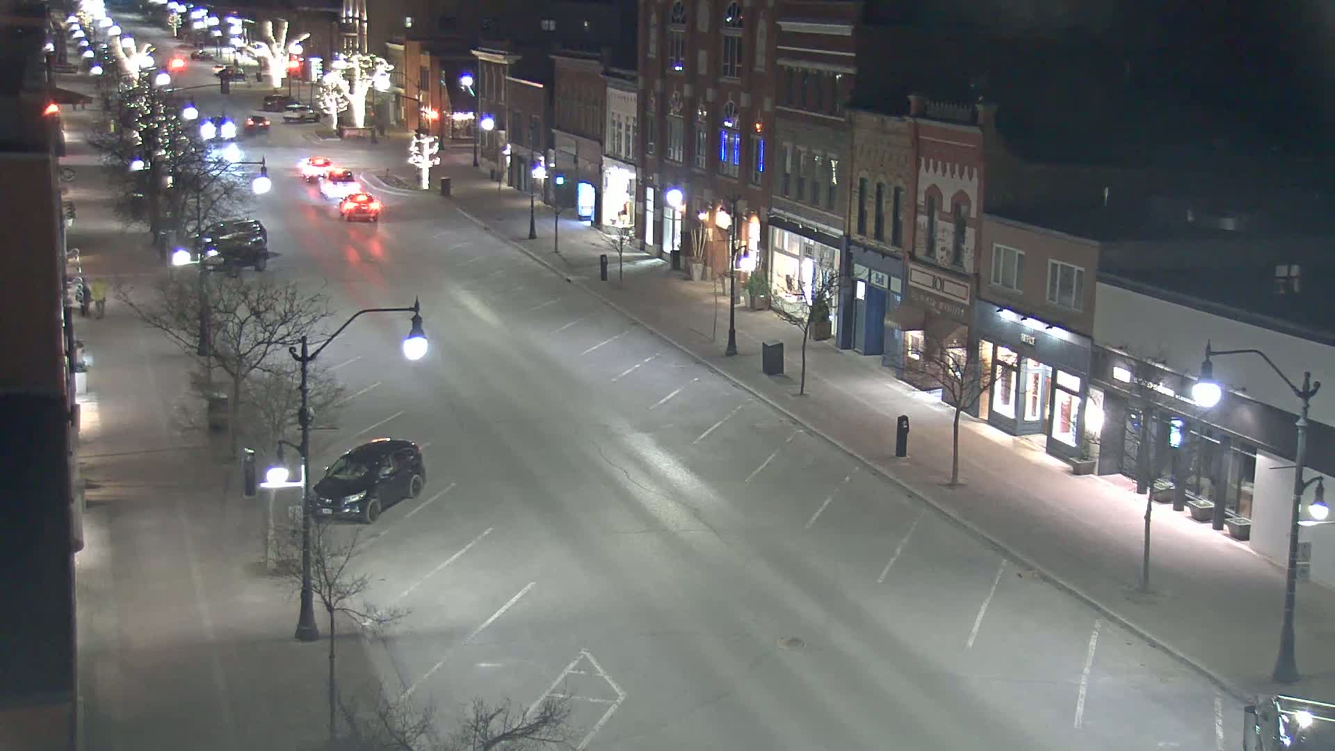 A mostly empty street at night, lined with shops and streetlights, has a few cars driving and parked along the curb.