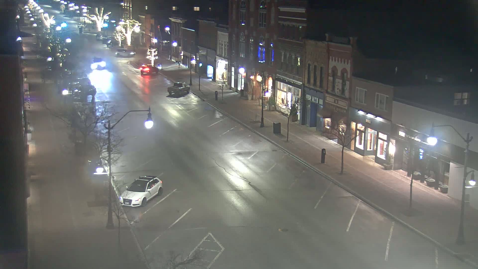 A mostly empty, wet street at night lined with parked cars and shops, lit by streetlights.