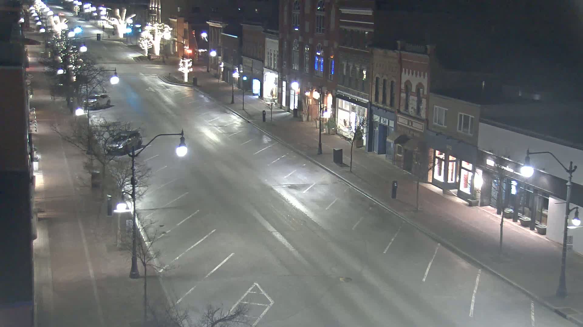 A mostly empty street at night, lined with brick buildings and streetlights, is lightly wet from a recent rain or snow.