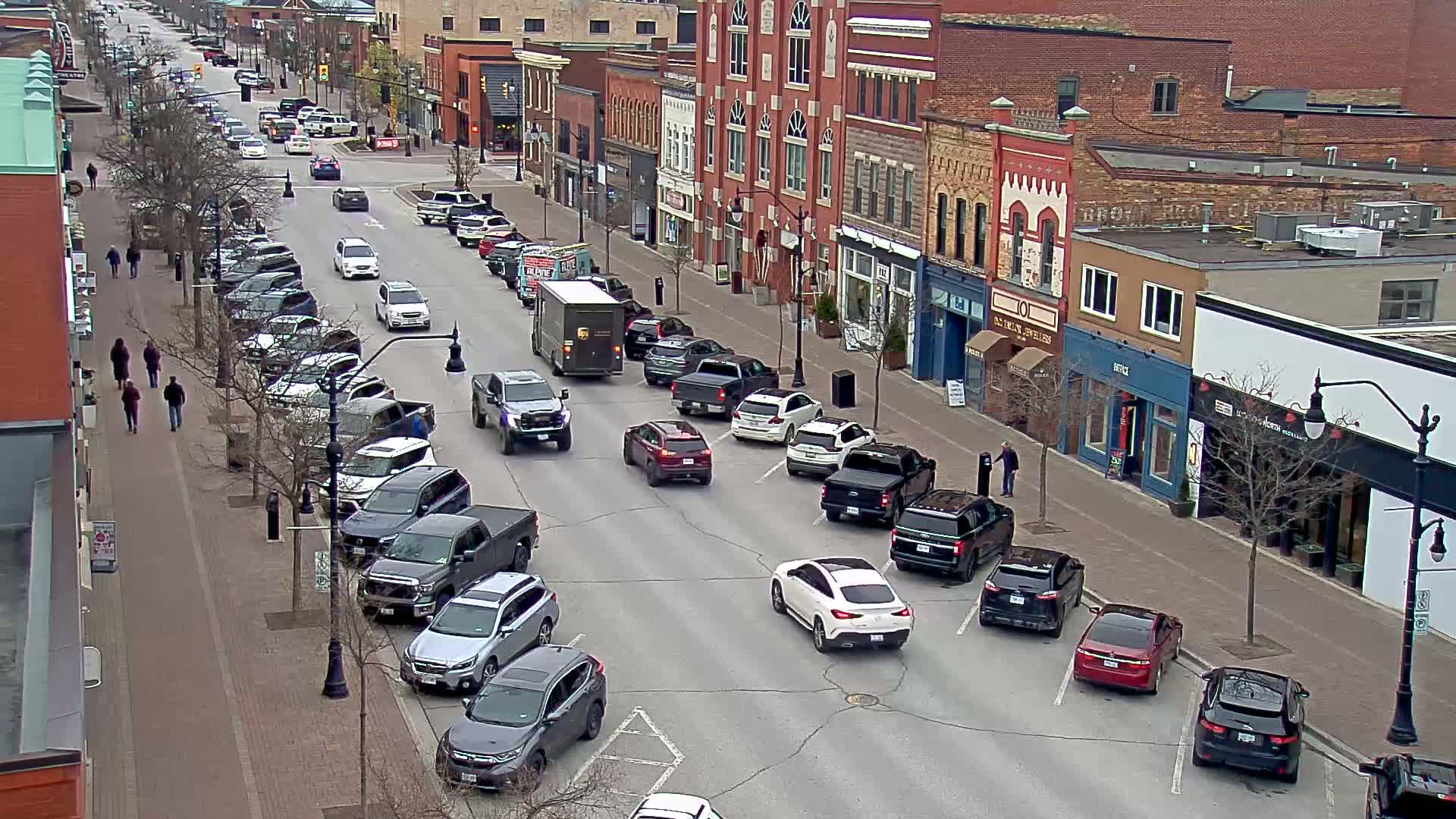 A street lined with brick buildings and parked cars on a cloudy day, with pedestrians walking on the sidewalk and several vehicles moving along the road.