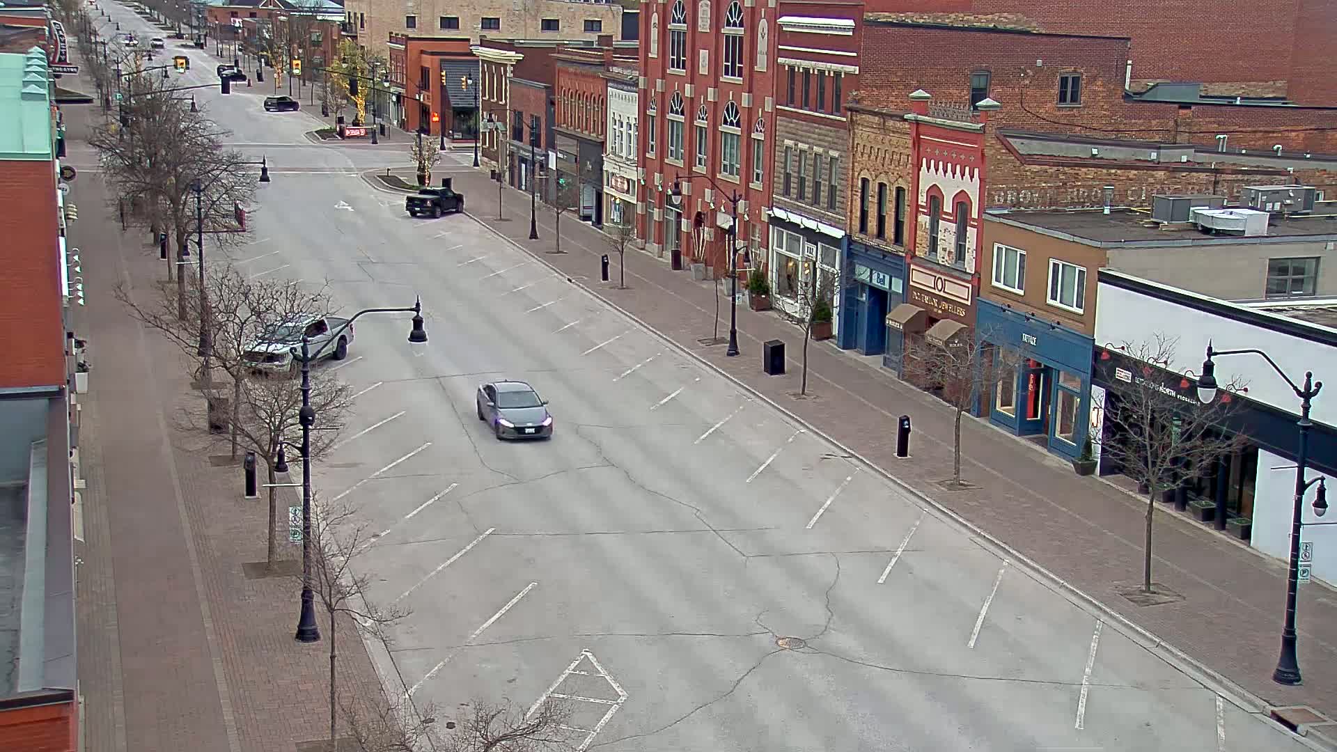 A mostly empty street with parked cars and a few driving cars lines brick buildings under a cloudy sky.