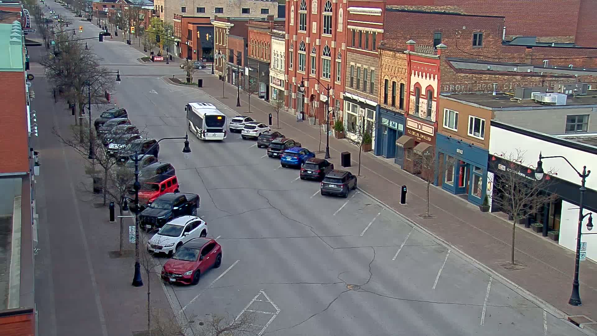 A mostly empty street lined with brick buildings and parked cars, a bus is stopped in the road, under a partly cloudy sky.