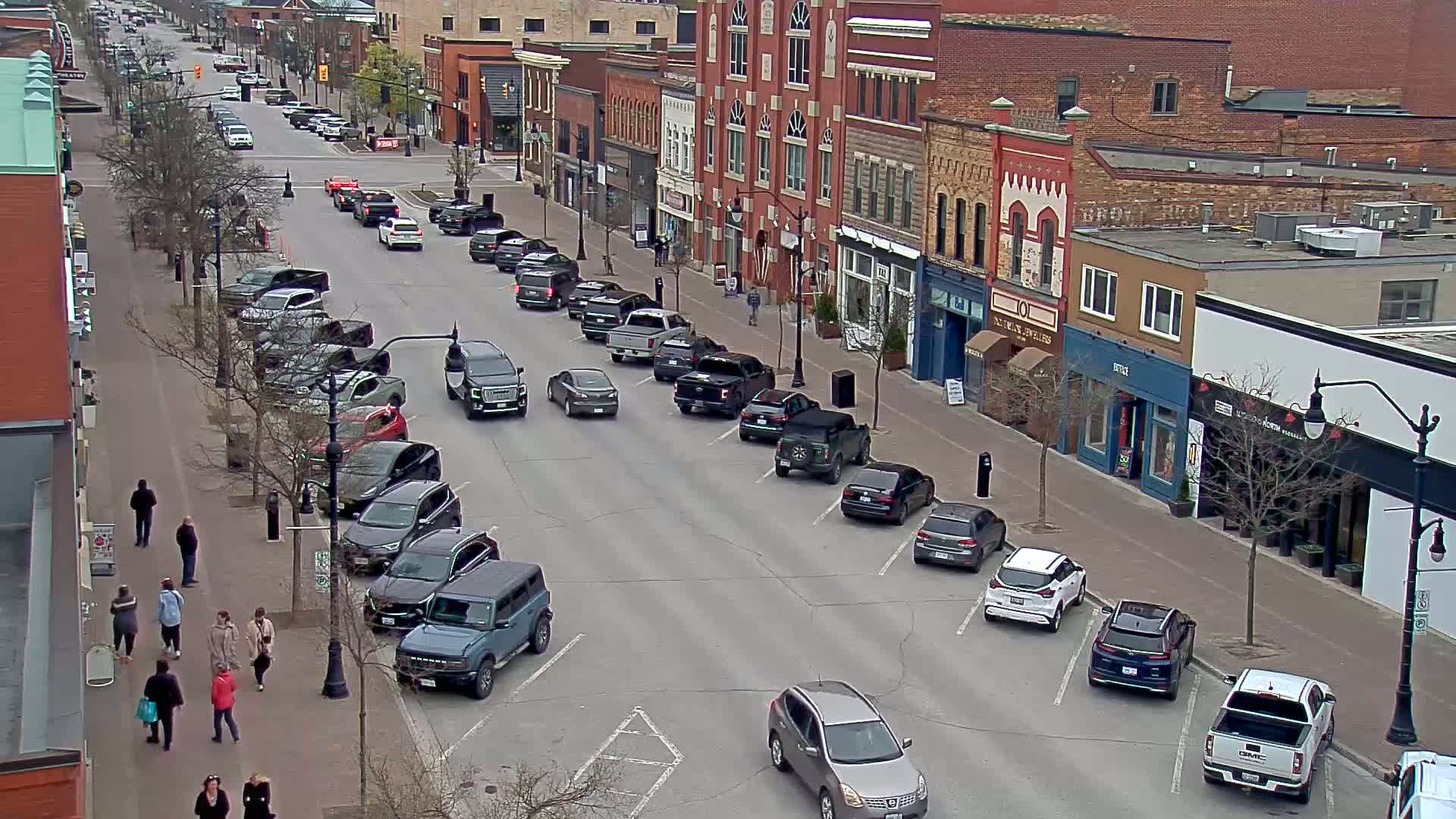 A street lined with parked cars and pedestrians, flanked by brick buildings on a partly cloudy day.