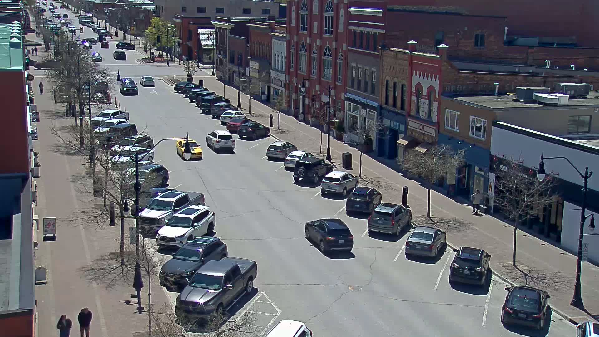 A sunny day shows a city street lined with parked cars, alongside brick buildings with shops on the ground floor.