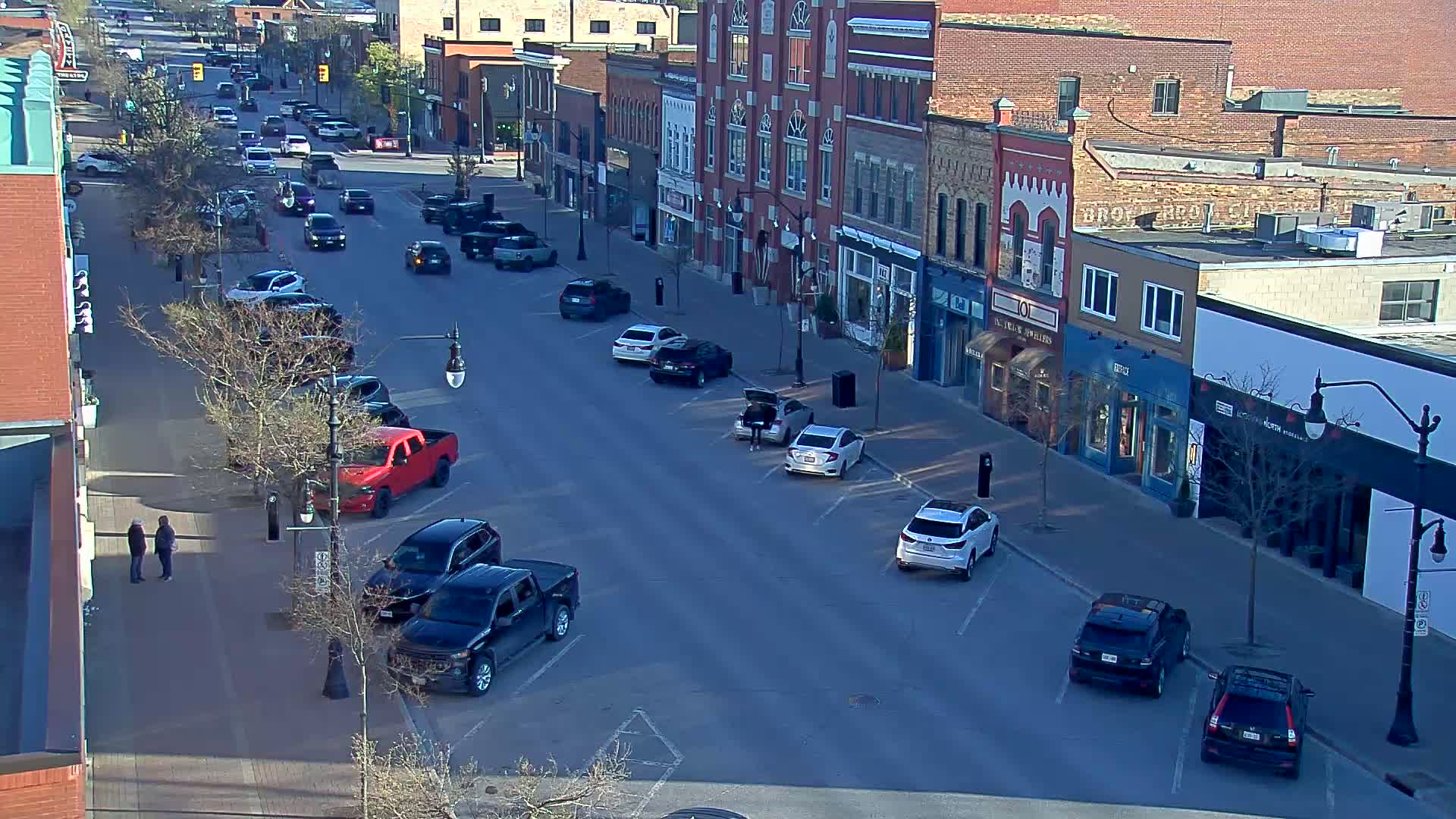 A sunny day reveals a mostly empty street lined with brick buildings and parked cars, with a few pedestrians visible.