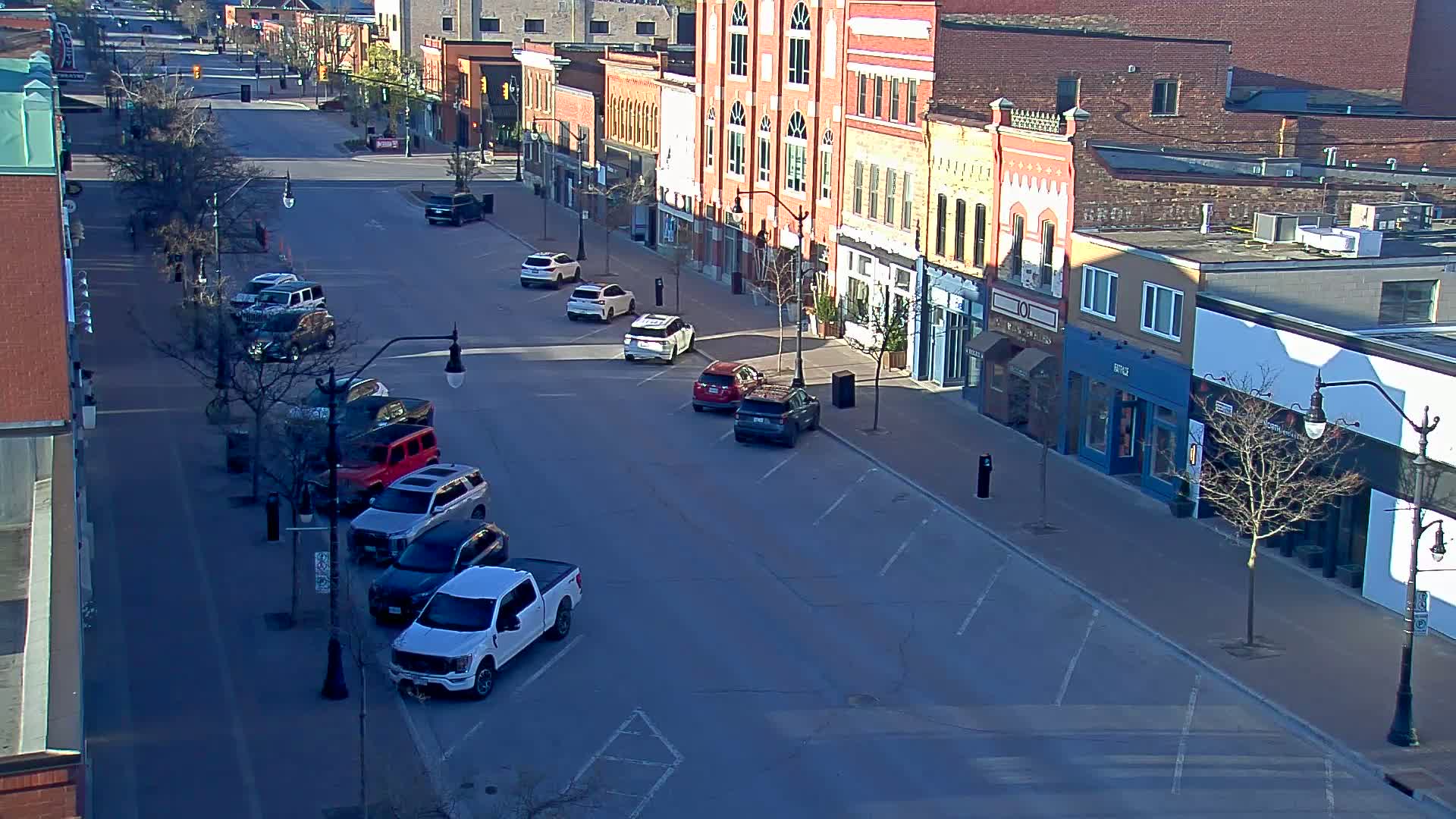 A mostly empty street lined with brick buildings on a sunny day, several cars parked along the sides.