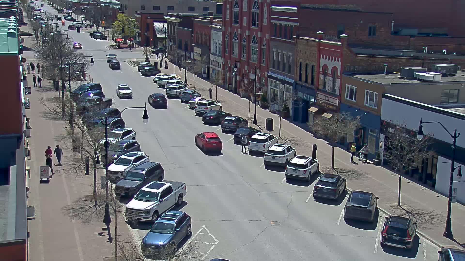 A sunny day on a city street with cars parked along both sides and a few people walking on the sidewalk.