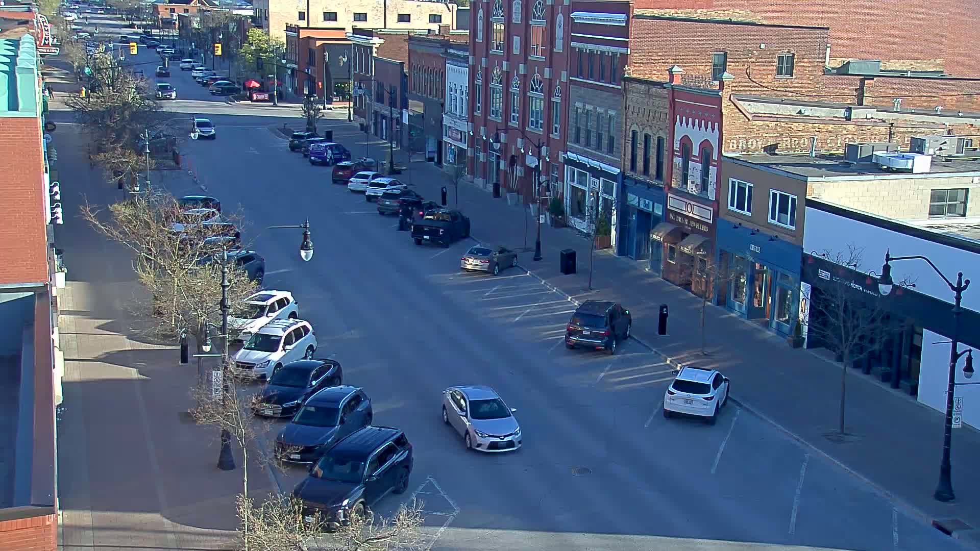 A sunny street scene shows several parked cars along a row of brick buildings, with a few cars driving on the road.