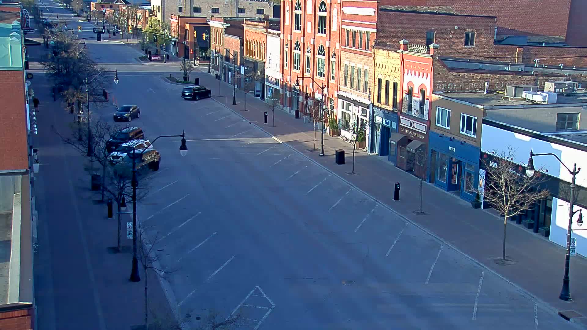 A mostly empty street lined with brick buildings on a sunny day, with a few cars parked and driving.