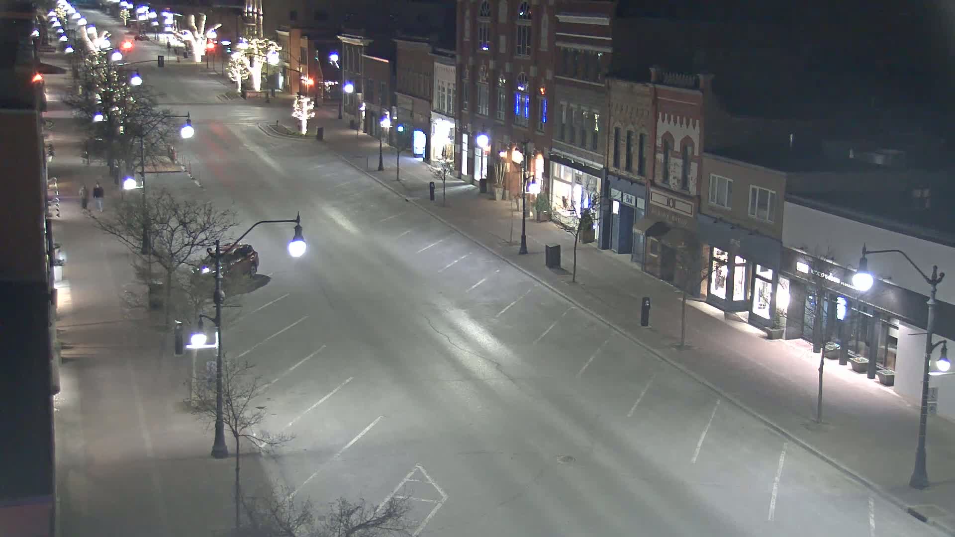 A mostly empty street at night, lined with shops and streetlights, is lightly dusted with snow.