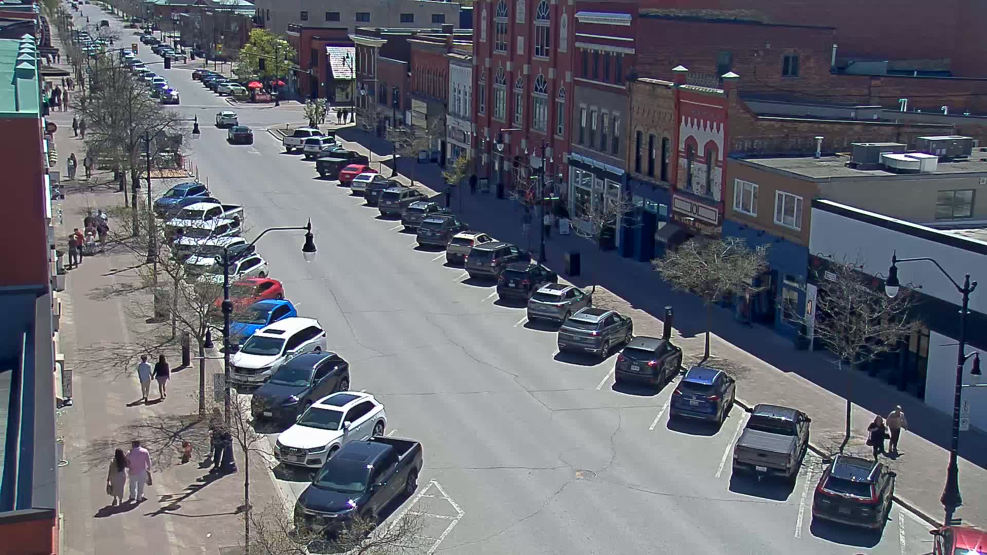 A sunny street scene shows pedestrians strolling along a sidewalk lined with parked cars in front of brick buildings.