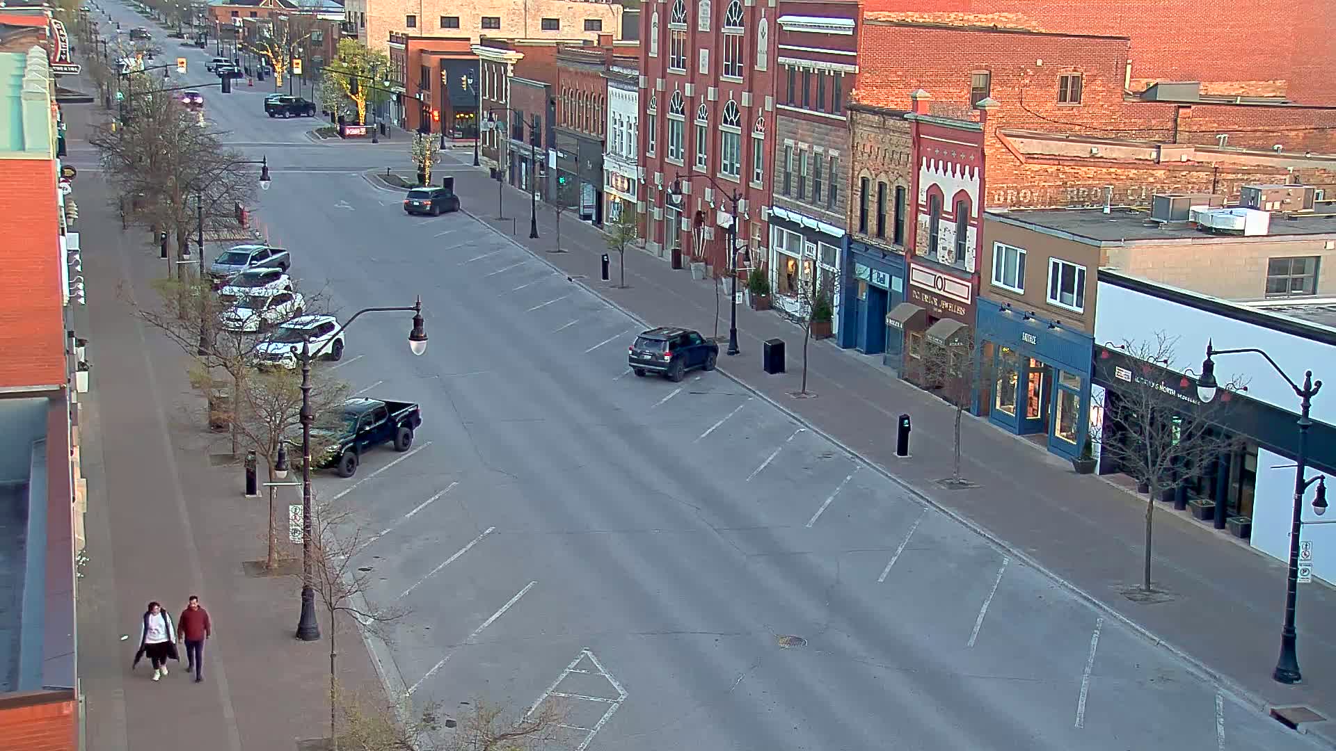 A mostly empty street lined with brick buildings and parked cars, with two pedestrians walking on the sidewalk under a partly sunny sky.