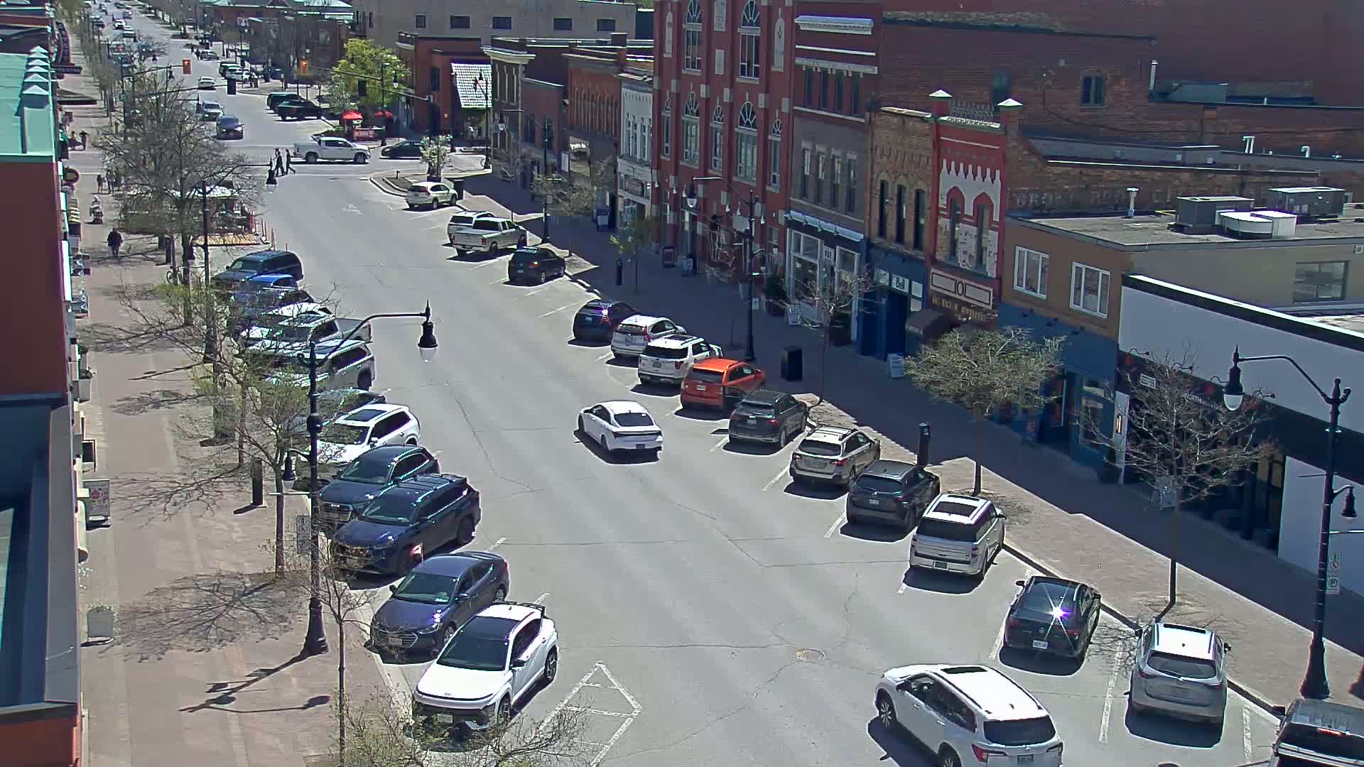 A sunny street scene shows cars parked on both sides of a road lined with brick buildings, and a few pedestrians walking on the sidewalk.