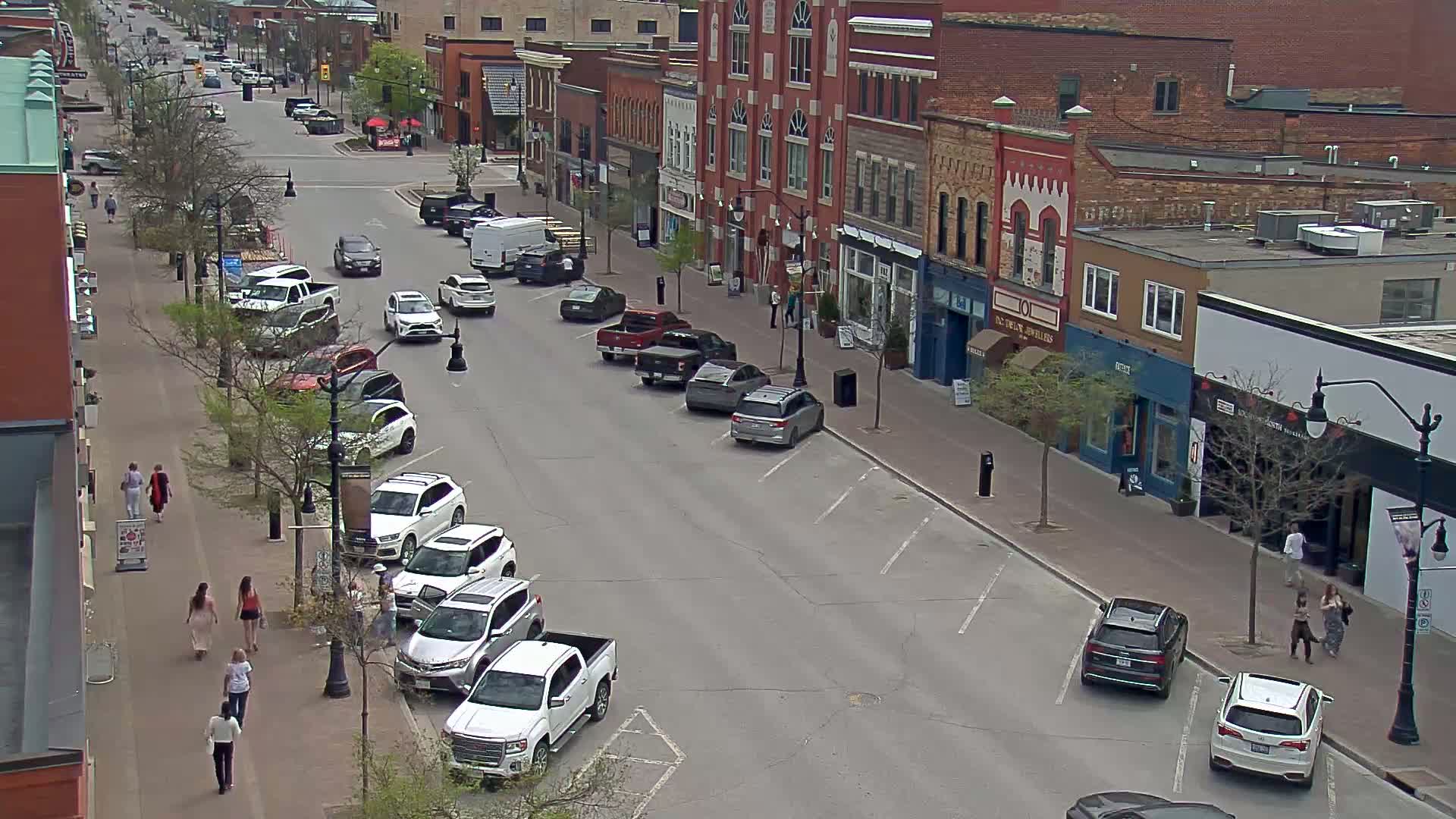 A street scene on a partly cloudy day shows several cars parked along the sides of a street lined with brick buildings and pedestrians walking on the sidewalk.