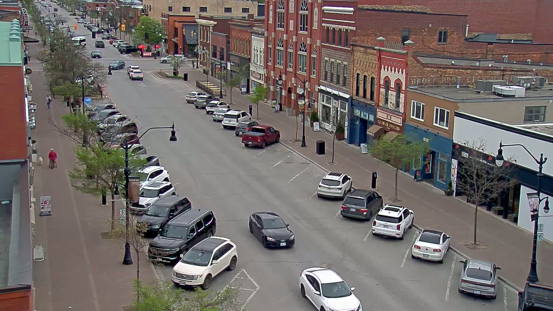 A mostly sunny street scene shows cars parked along the sides of a two-lane road lined with brick buildings and leafy trees.