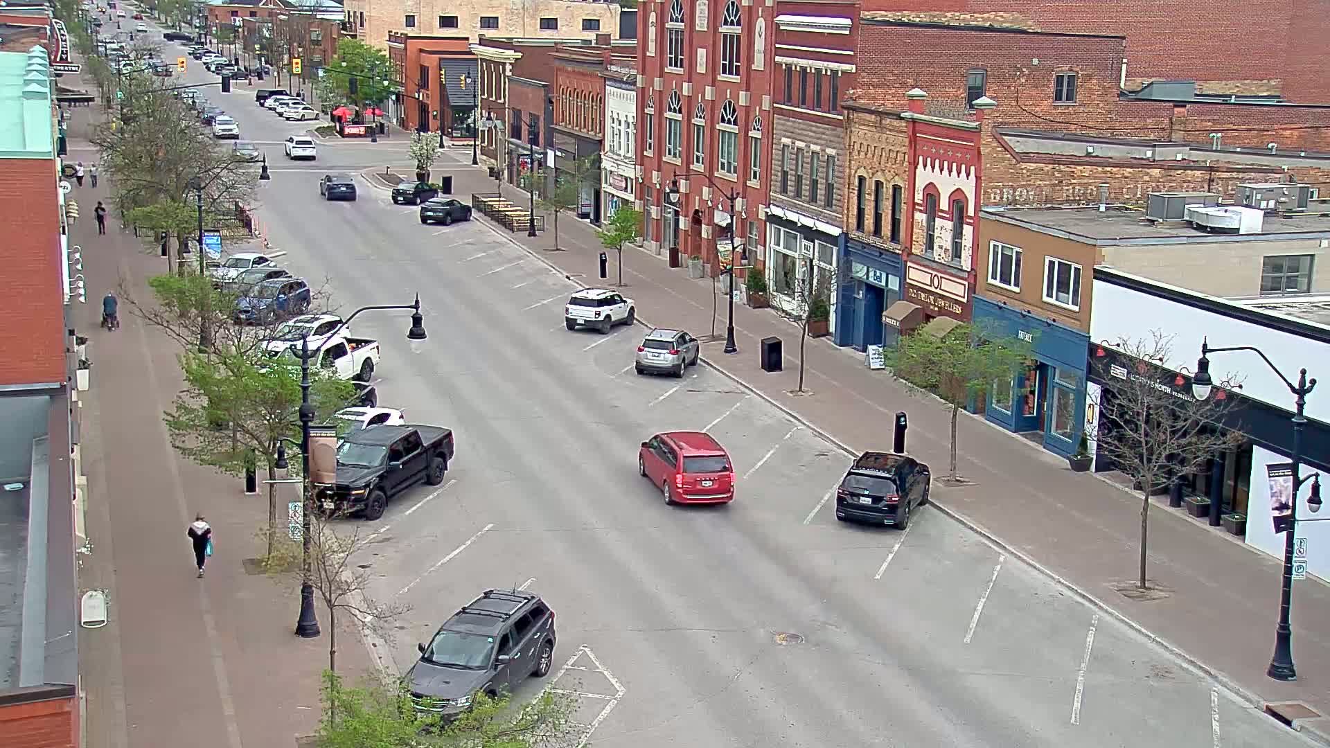 A mostly empty street lined with brick buildings on a partly sunny day shows several parked cars and a few pedestrians.