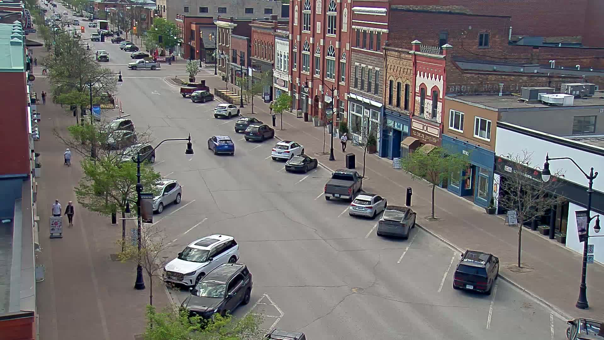 A street lined with parked cars and brick buildings on a sunny day, with a few pedestrians walking on the sidewalk.