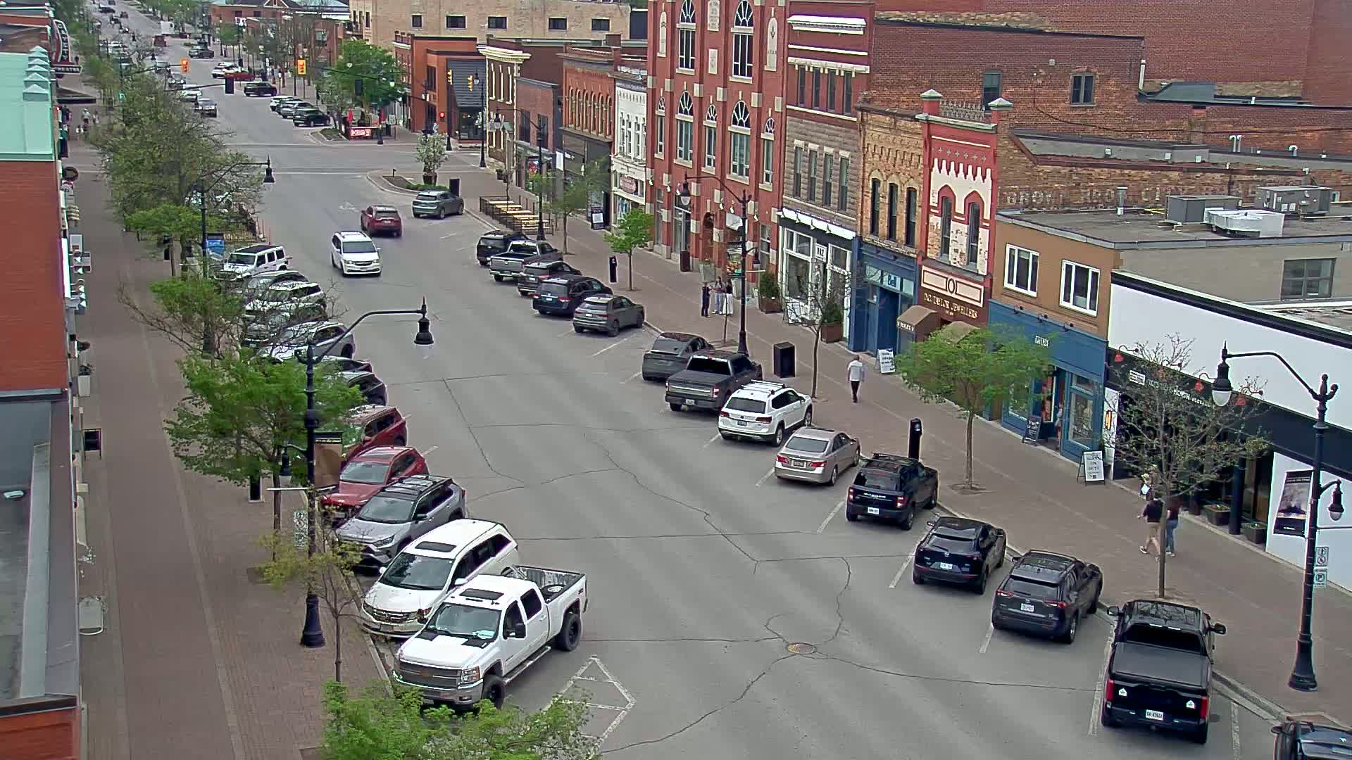 A street lined with parked cars and brick buildings, on a partly sunny day with green trees.