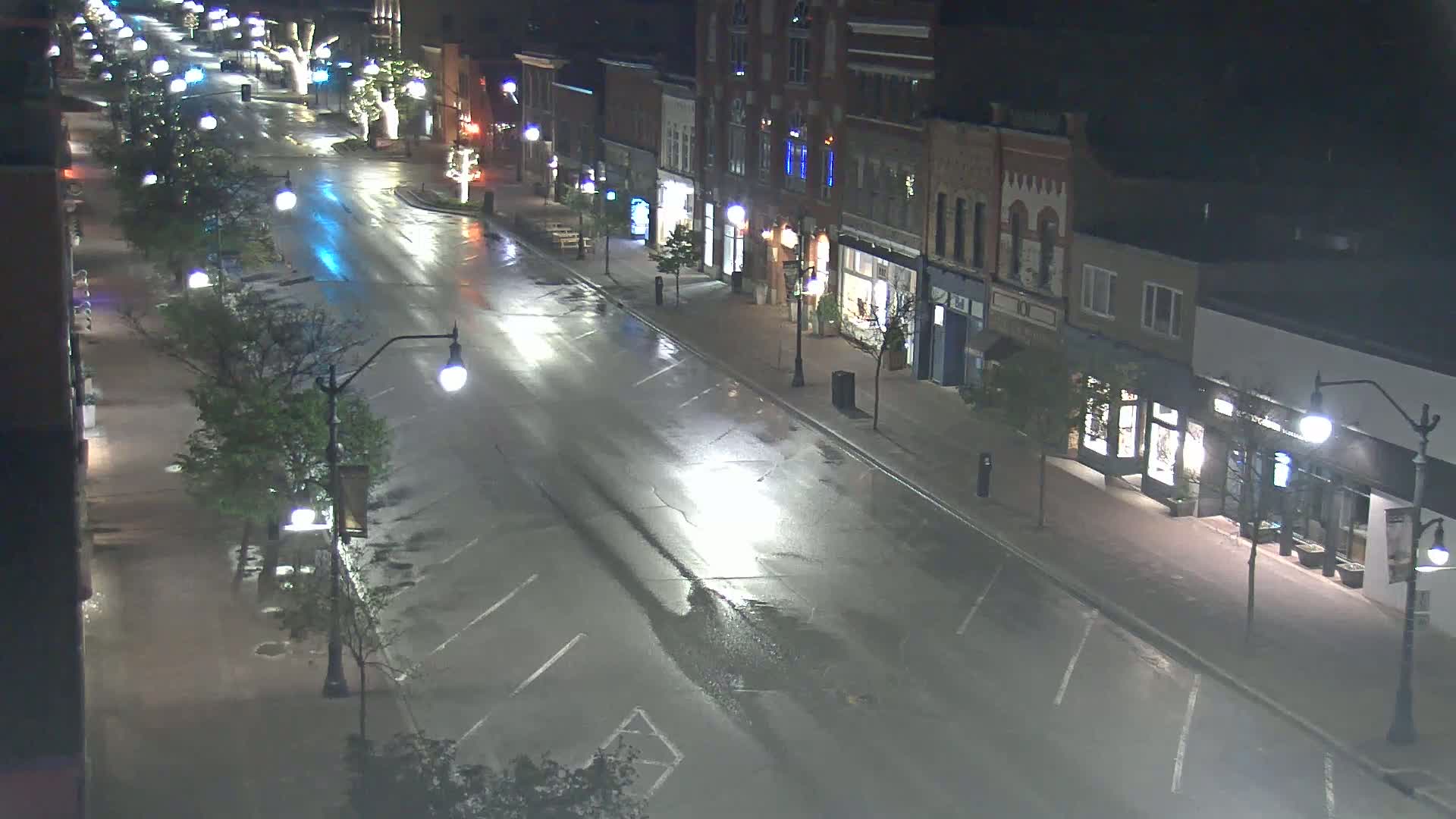 A wet, empty street at night, lined with multi-story buildings and streetlights, shows reflections of light on the road.