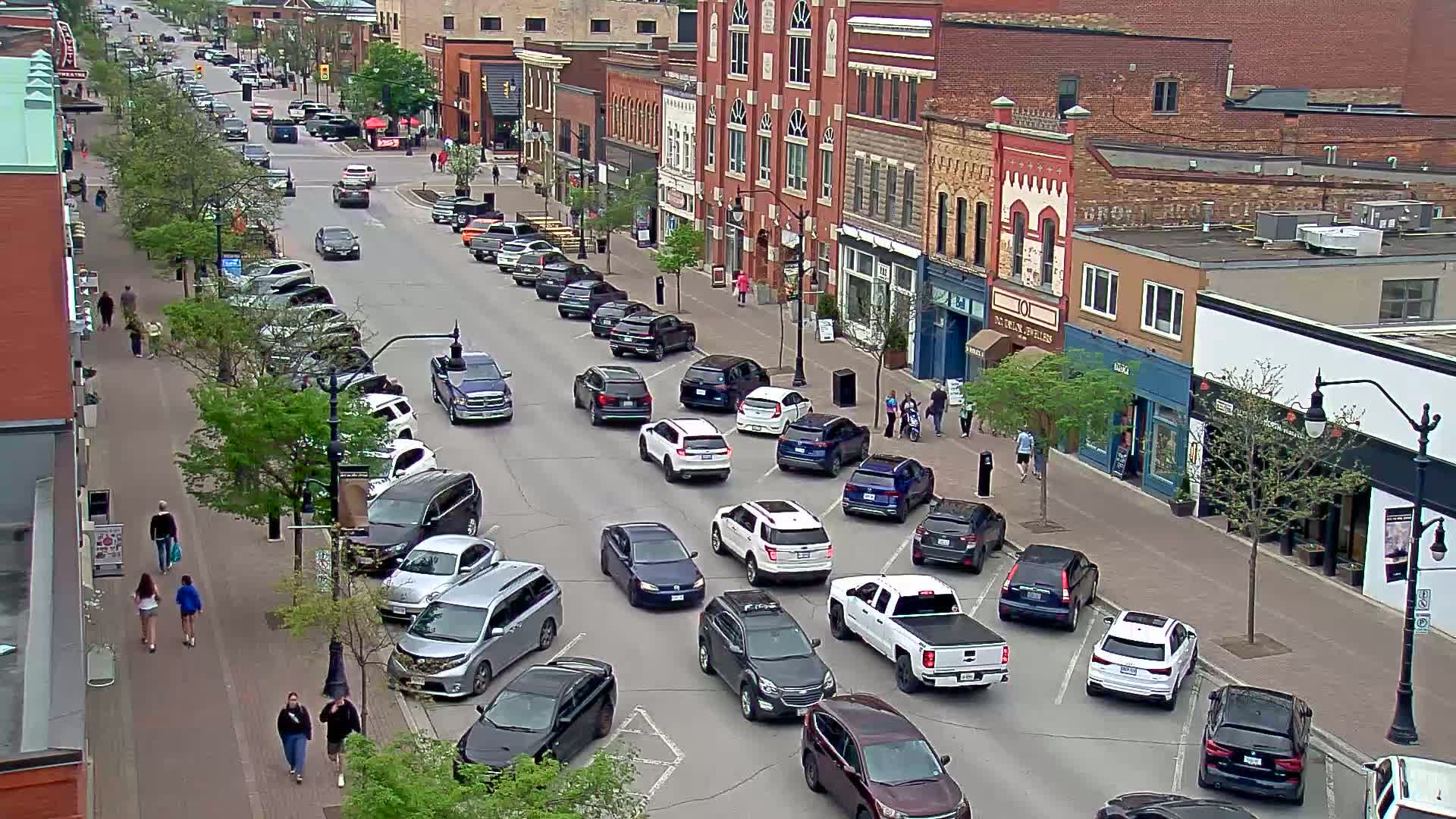 A partly sunny day reveals a busy street scene in a downtown area with cars parked on both sides of the street and several vehicles driving, while pedestrians stroll along the sidewalks past brick buildings.