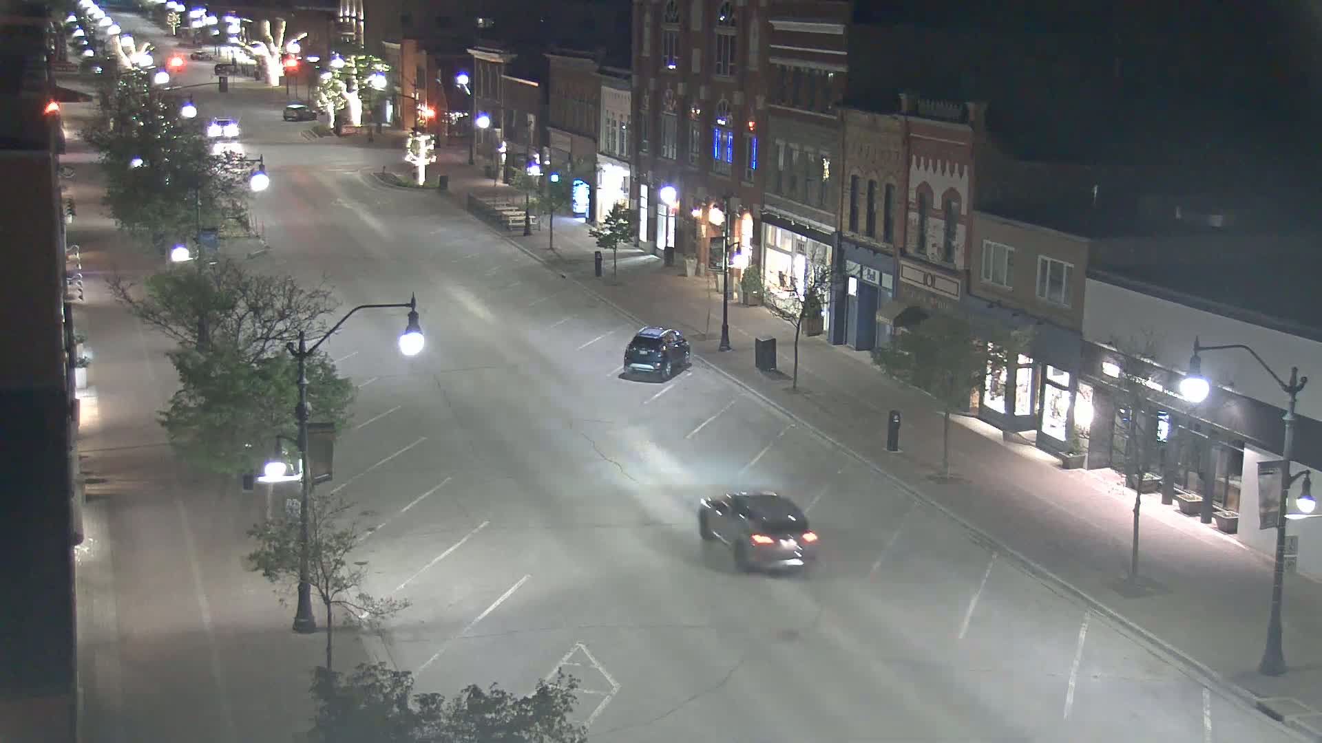 A mostly empty street at night lined with buildings and trees, illuminated by streetlights, with two cars driving on the road.