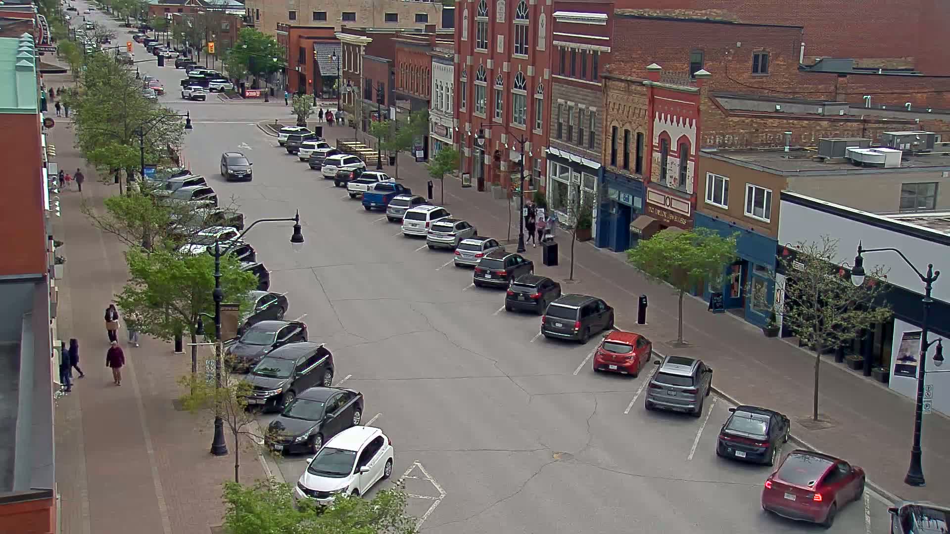 A street lined with parked cars and brick buildings, with pedestrians walking on the sidewalk under a partly sunny sky.