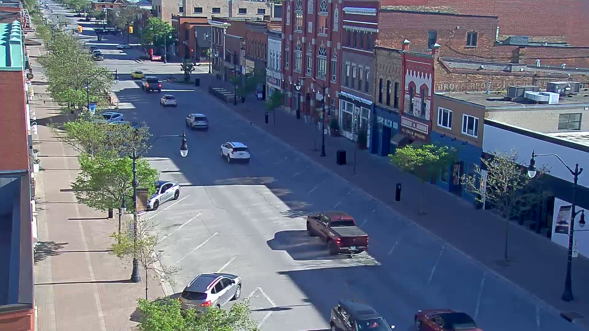 A sunny street scene shows several cars driving on a city street lined with brick buildings and young leafy trees.