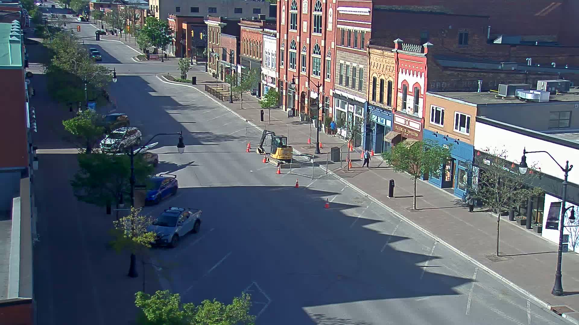 A mostly empty street lined with brick buildings on a sunny day, with a small excavator and construction cones in the middle of the road.