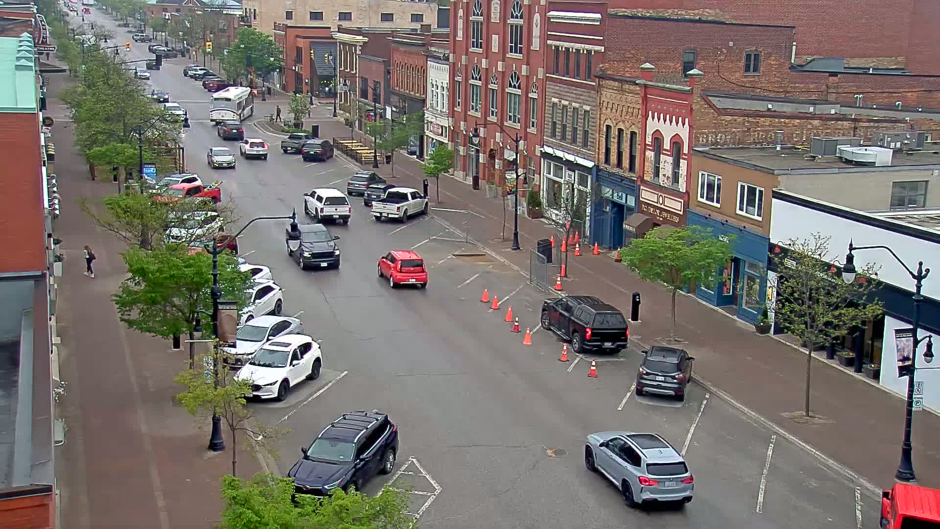 A street scene on an overcast day shows several cars parked and driving along a city block with two and three-story brick buildings lining both sides of the road, many with ground-floor businesses.