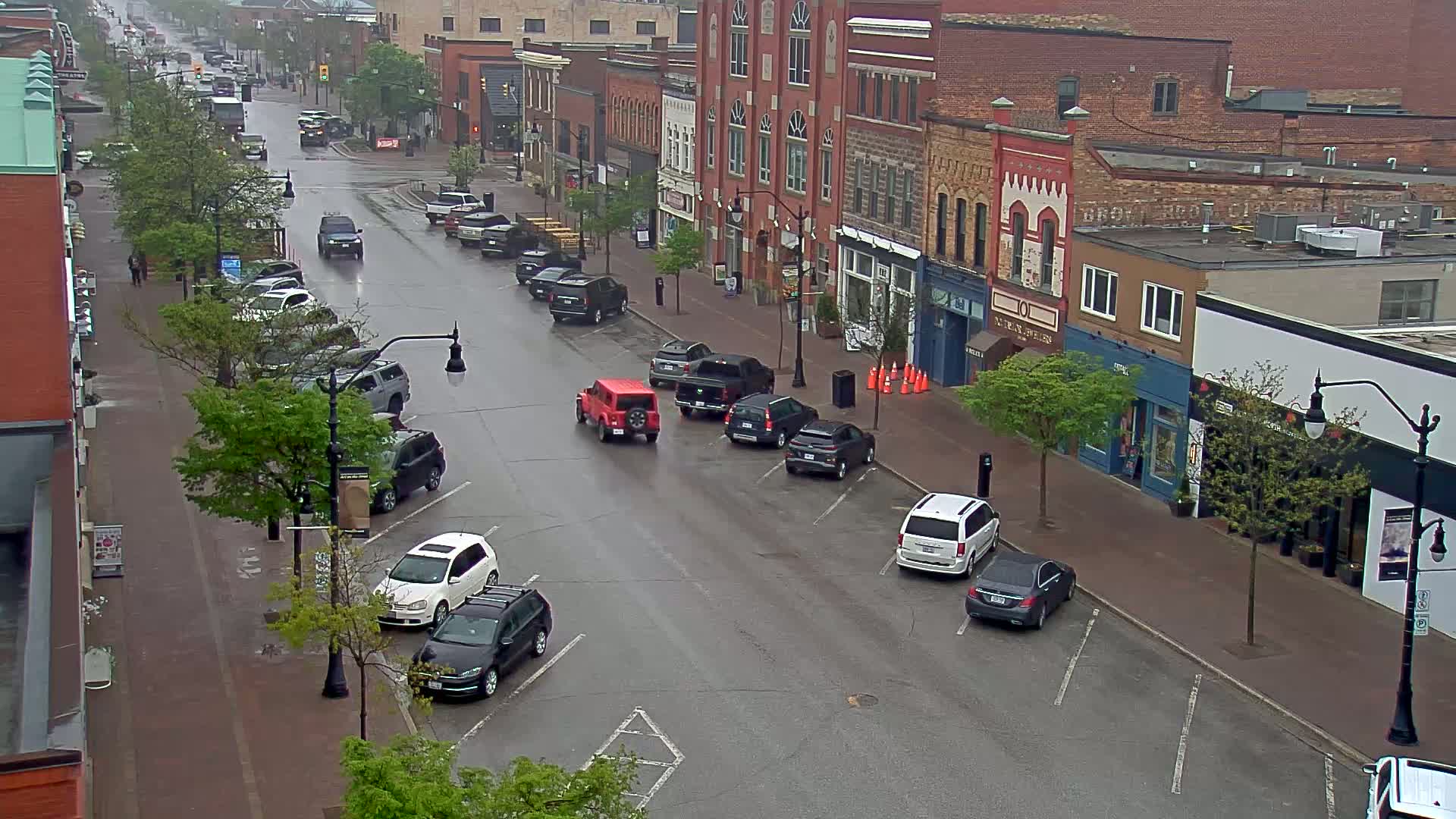 A rain-slicked street lined with parked cars and brick buildings, with a few cars driving down the middle of the road.