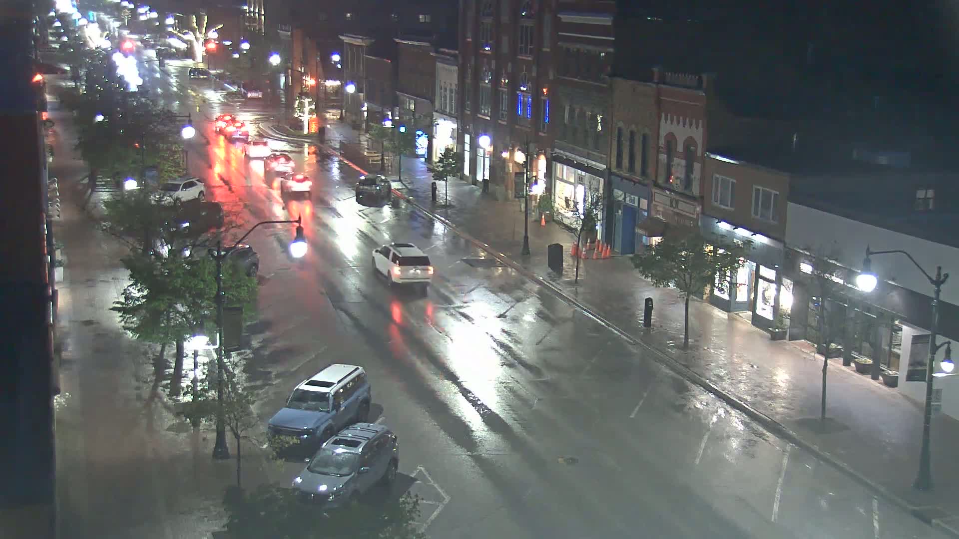 A rain-slicked street at night shows several cars driving and parked in front of brick buildings with storefronts.