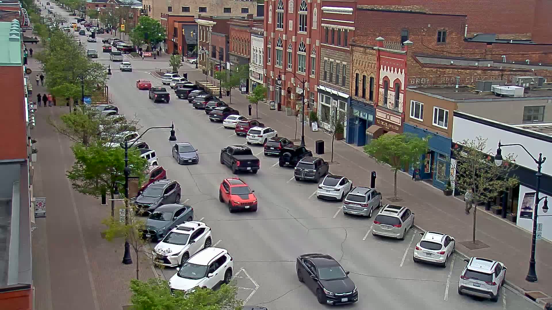 A street scene shows cars parked on both sides of a road lined with brick buildings under a partly sunny sky.