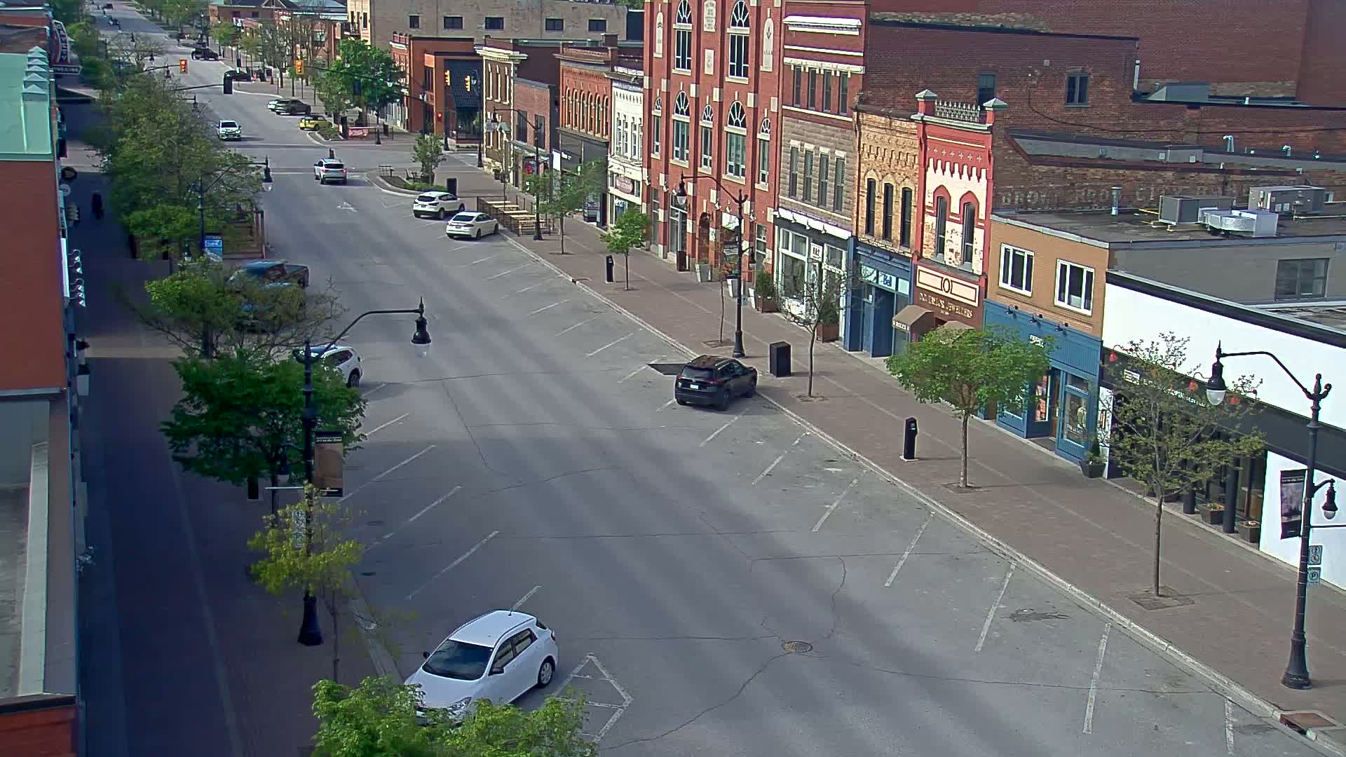 A mostly empty street lined with brick buildings and young trees, under a partly sunny sky.