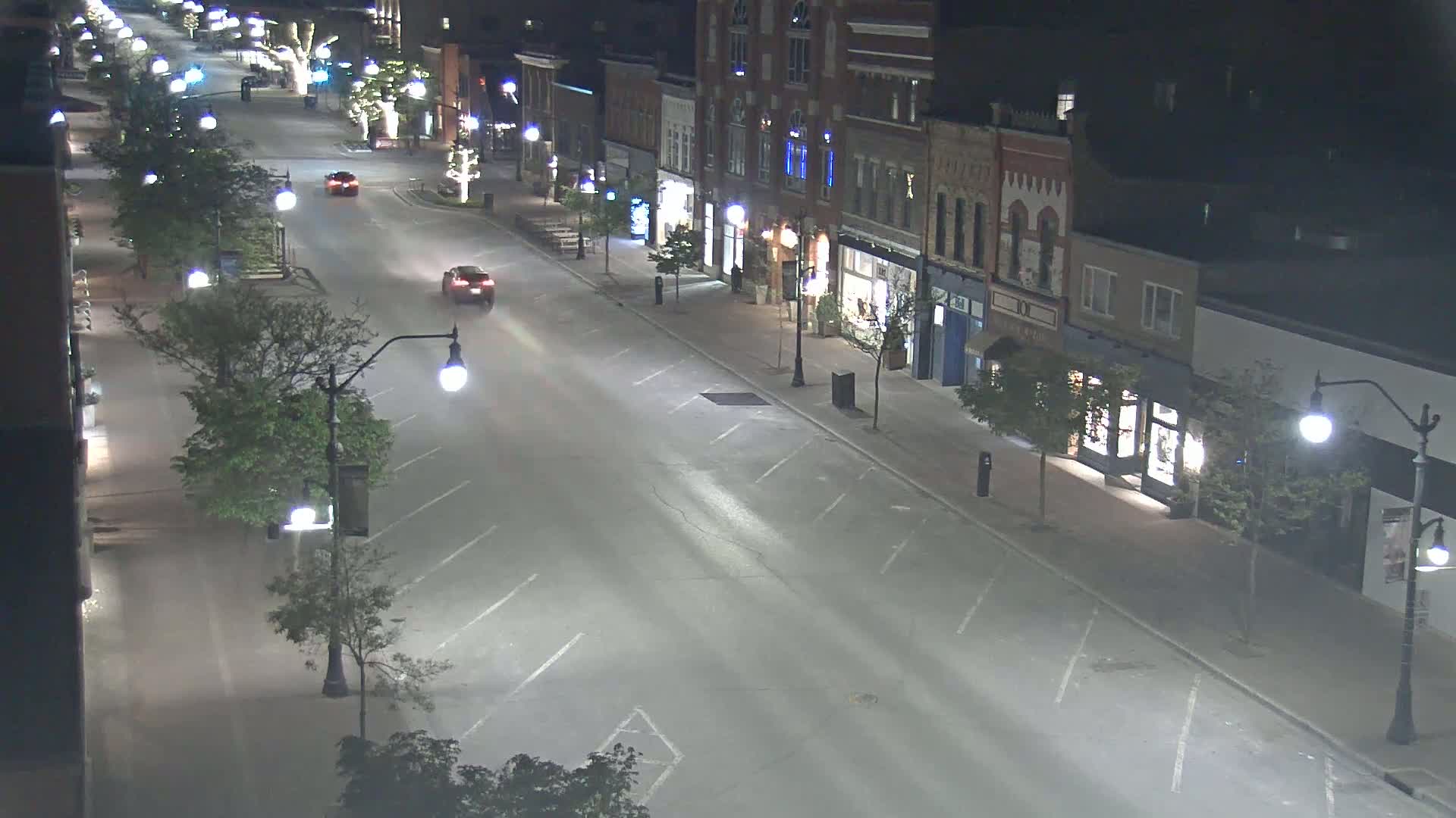 A mostly empty street at night, lined with shops and trees, is lit by streetlights.