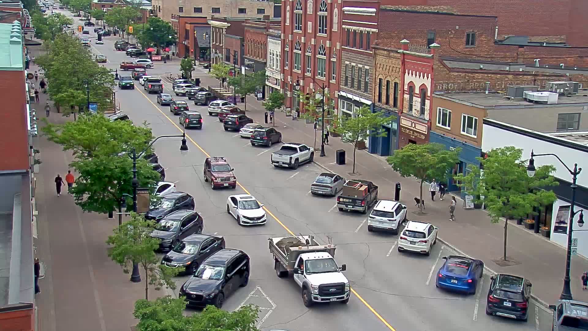 A street scene on a sunny day shows parked cars lining both sides of a roadway, with additional cars driving down the center, and pedestrians walking along the sidewalks in front of brick buildings.