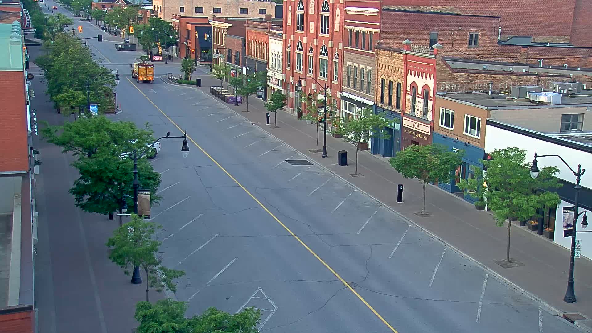 A mostly empty street lined with brick buildings and young green trees under a clear sky.