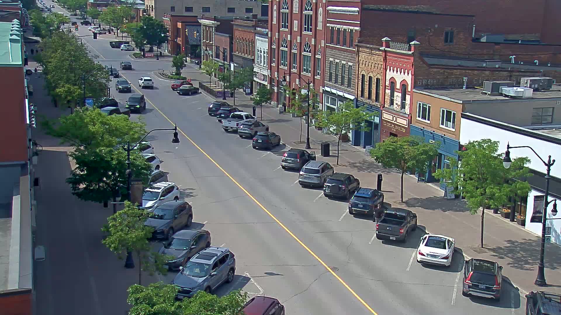 A sunny street scene shows cars parked on both sides of a road lined with two- and three-story brick buildings and leafy green trees.