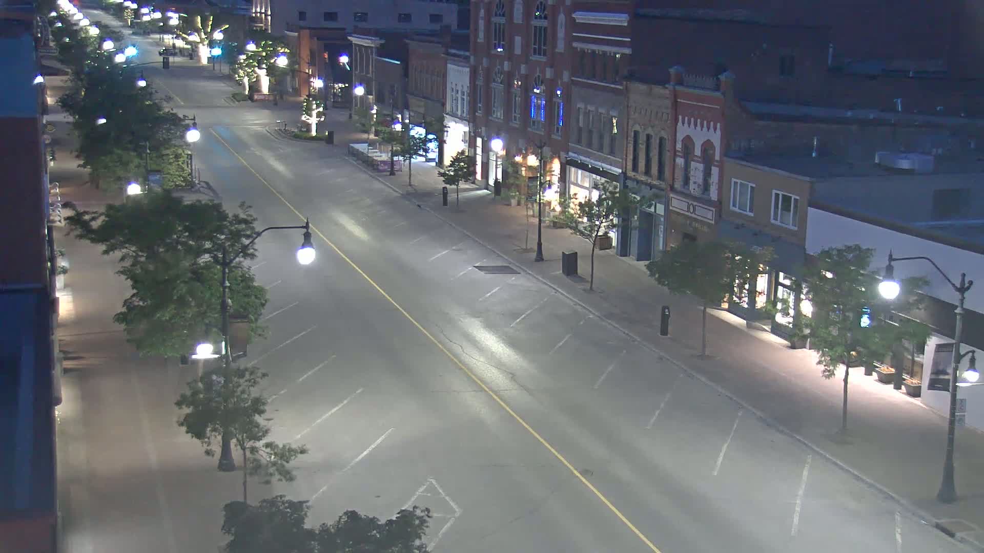 A mostly empty, well-lit street at night lined with shops and trees.