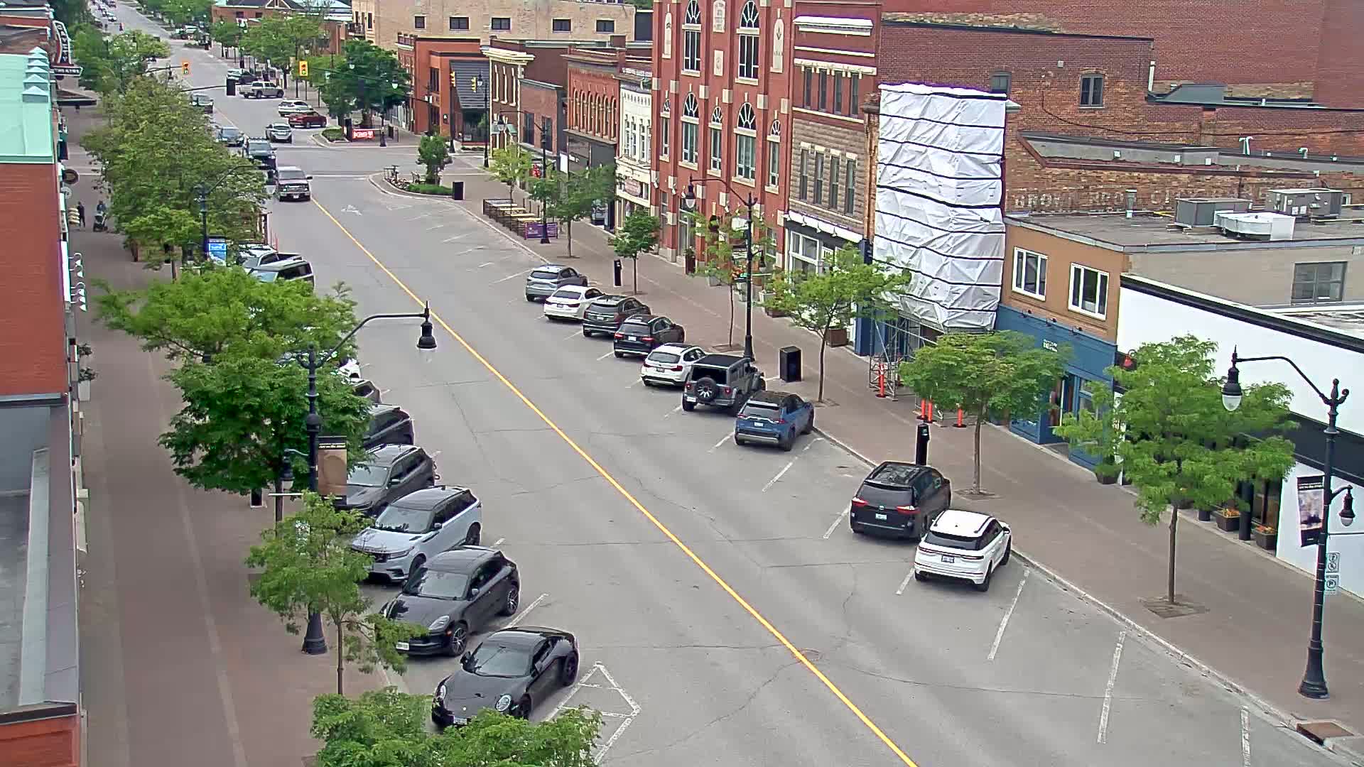 A city street scene on a partly sunny day shows several cars parked along the sides of a road, in front of brick buildings, some of which are under construction.